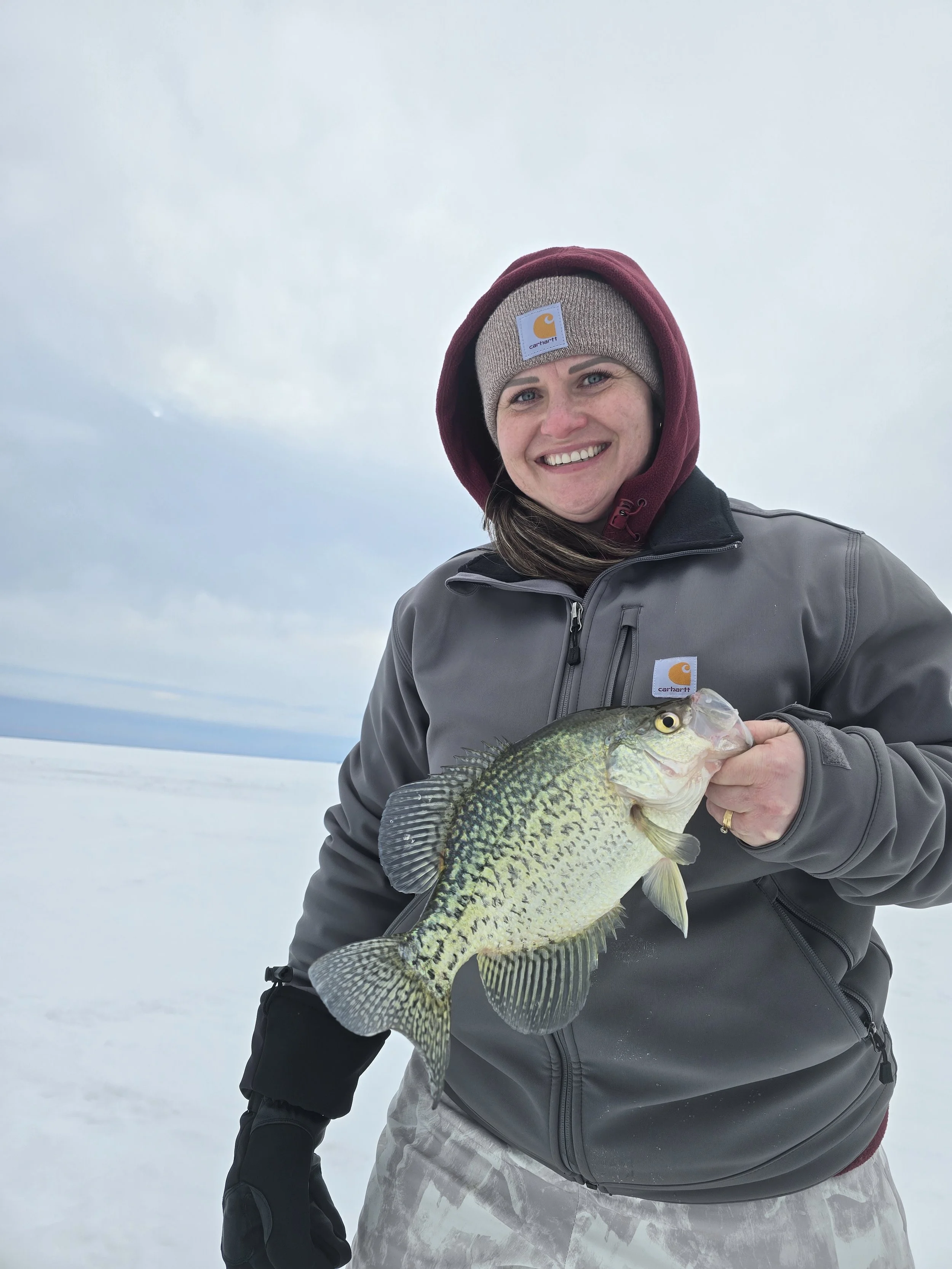 A smiling woman in winter clothing holding a large fish outdoors in a snowy landscape, wearing a Carhartt beanie and gray jacket.