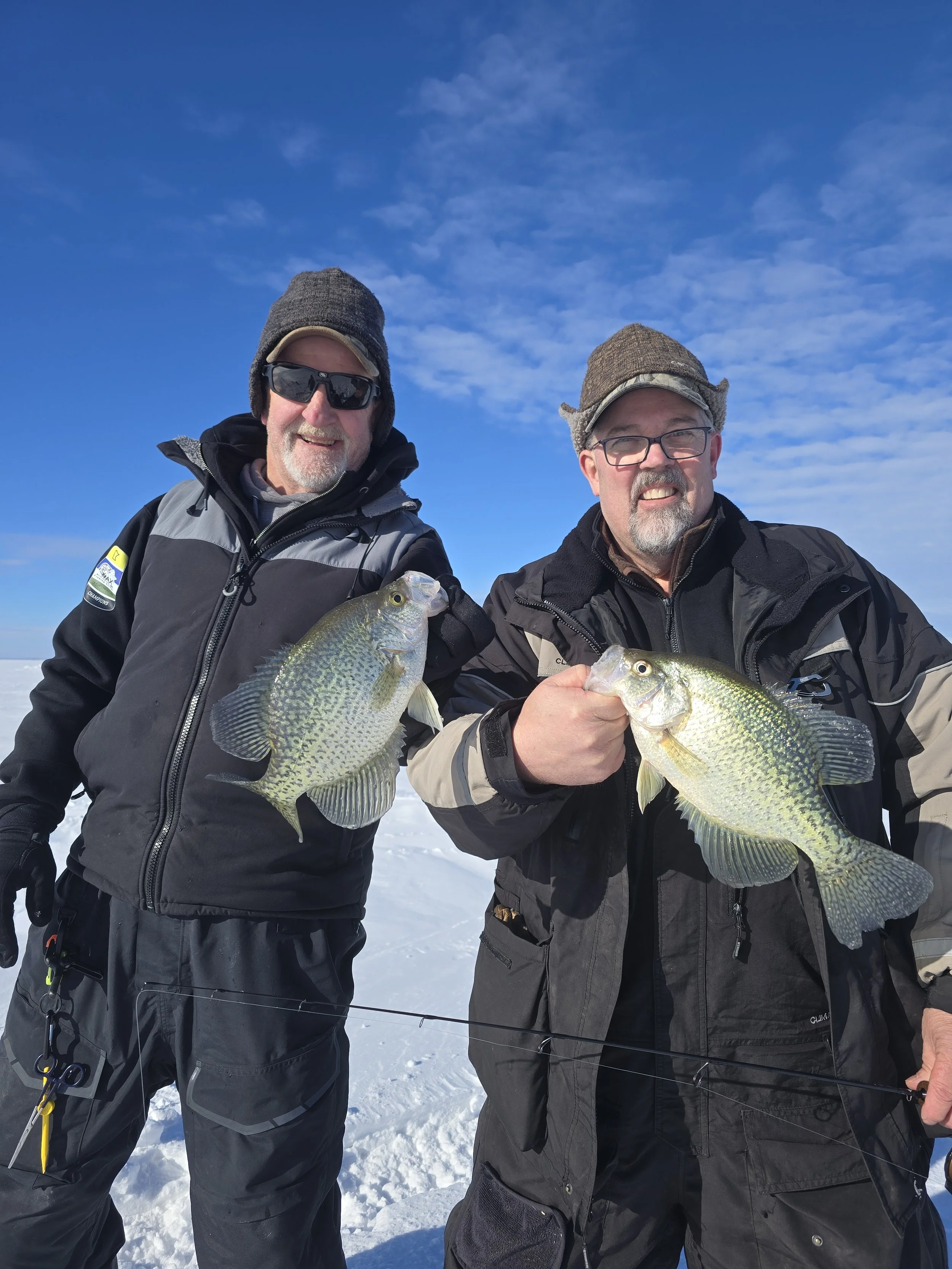Two men standing outdoors on snow, holding fish they caught, wearing winter gear and hats, with a blue sky and some clouds in the background.