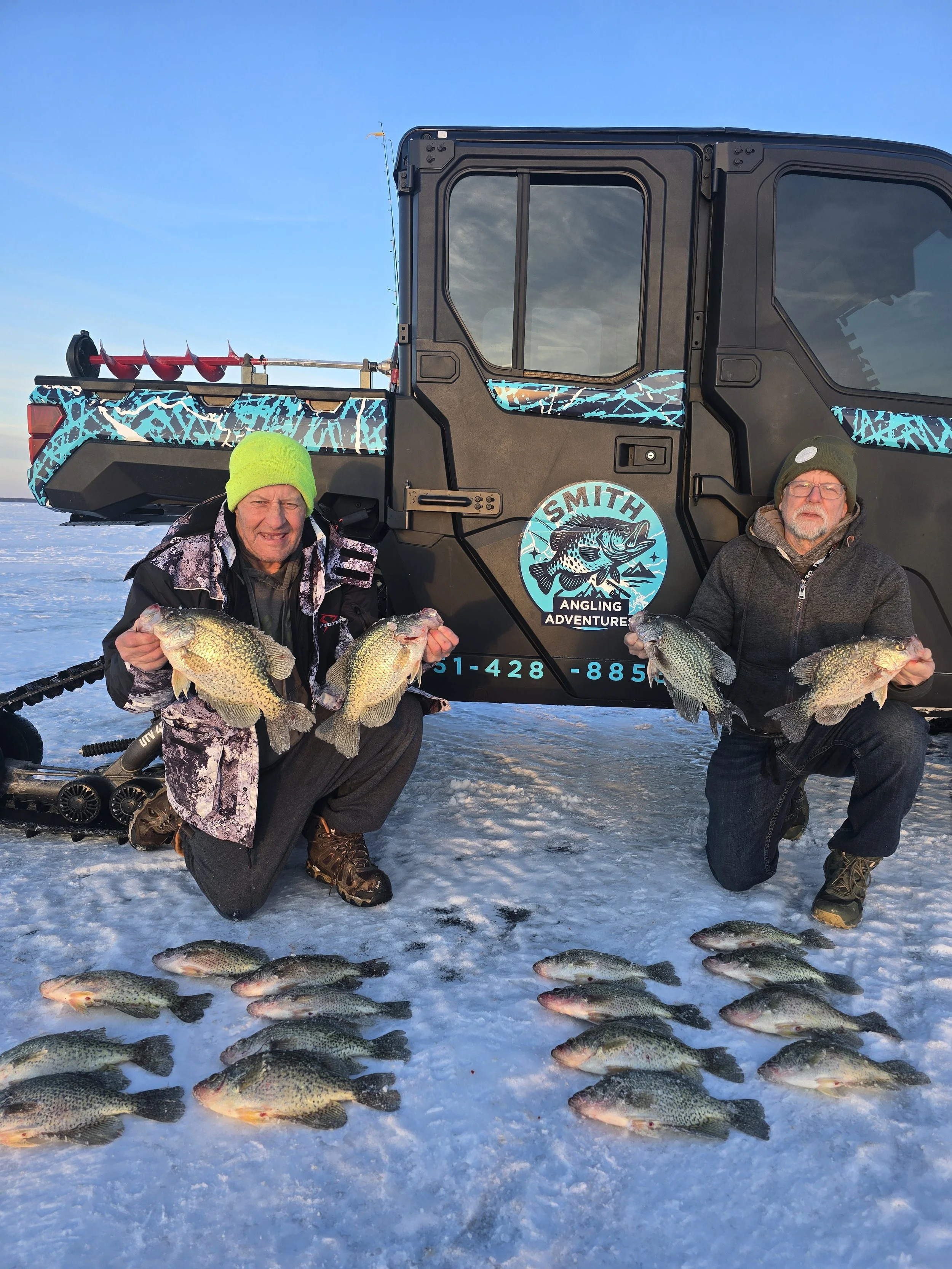 Two men kneeling on ice with a group of fish they caught, snow and a blue sky in the background, a black vehicle with a fishing-related logo and gear behind them.