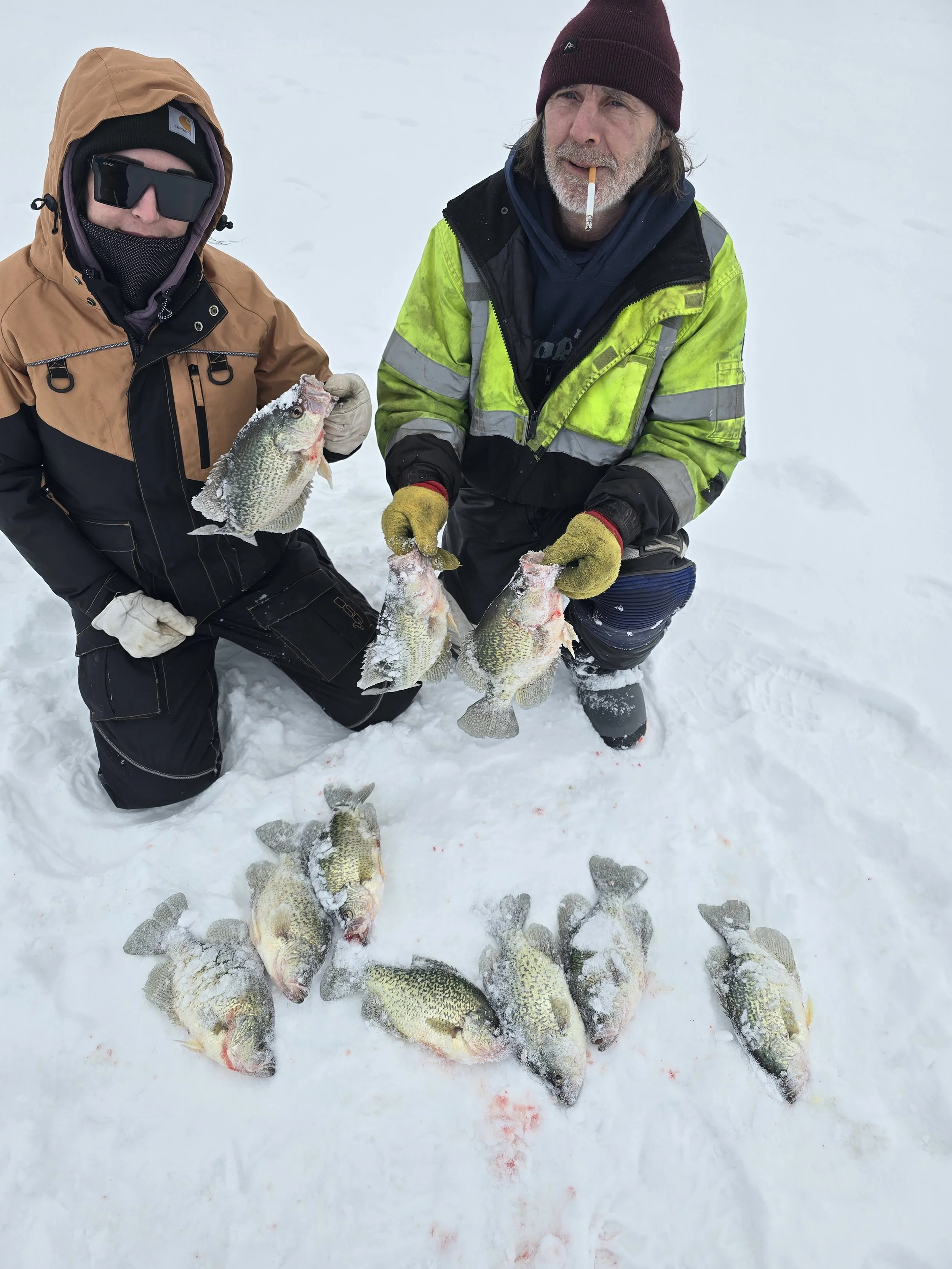 Two men dressed in winter clothing kneeling on snow-covered ground, displaying several caught fish. One man is holding a fish with ice on its scales, while the other is holding two fish, with more fish lying on the snow.