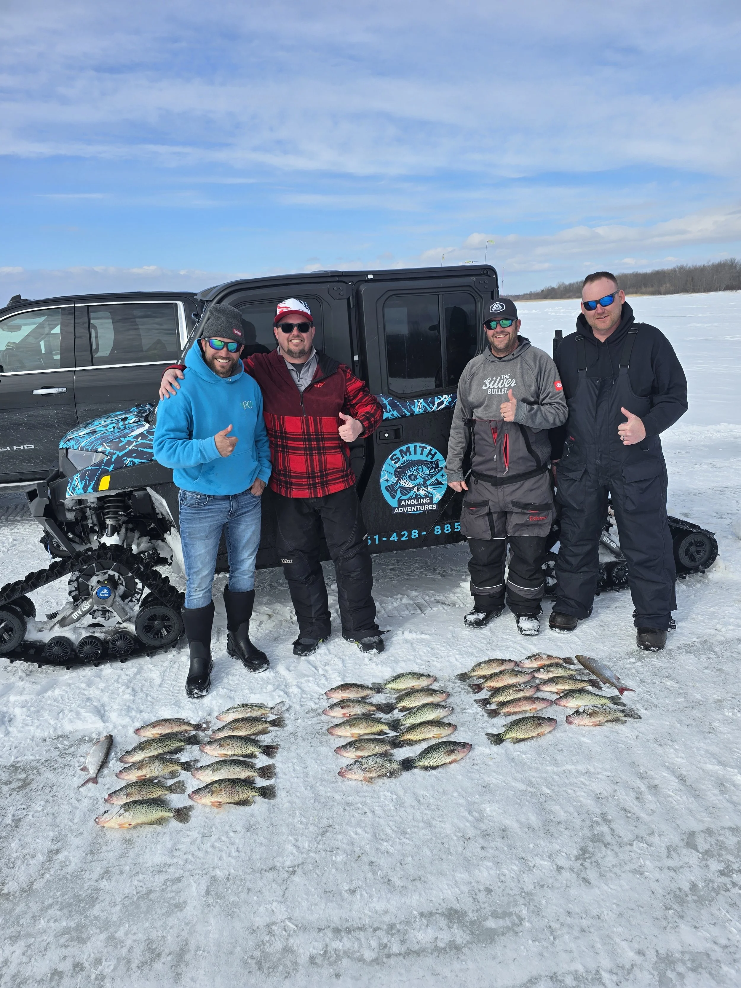 Four men in winter clothing and sunglasses standing on snow-covered ice, posing with a selection of fish they caught, with a black snowmobile and a black truck in the background and a snowy landscape under a cloudy sky.
