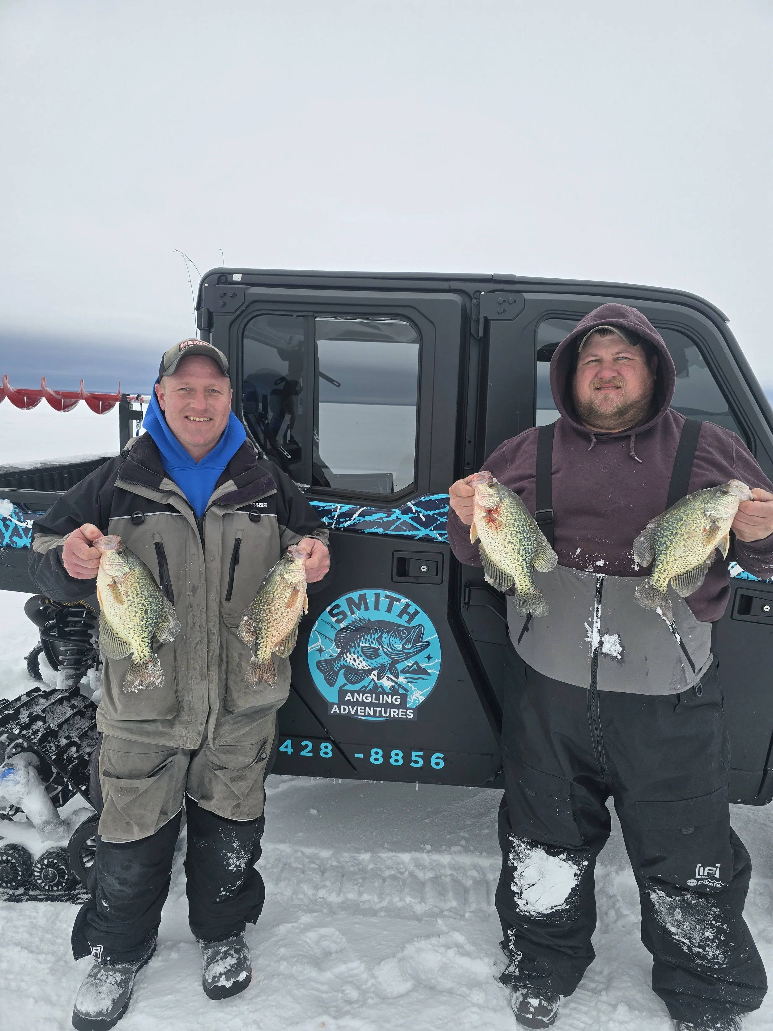 Two men in winter clothing holding large fish standing in front of a snow-covered vehicle with a logo that reads 'Smith Angling Adventures' and a phone number. They are on a snowy landscape with cloudy sky.