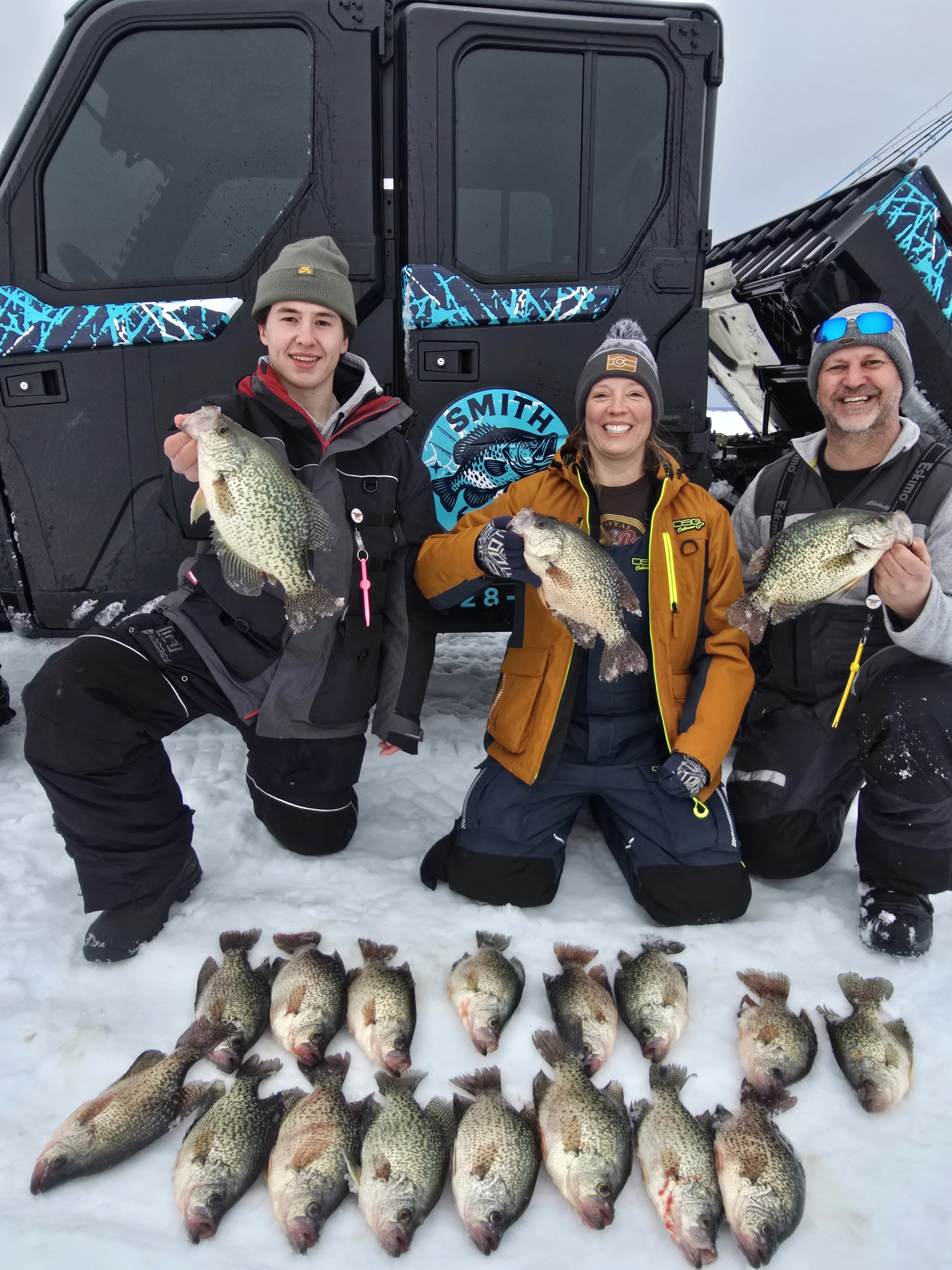 Three people kneeling on snow with a large catch of fish laid out in front of them and a snowmobile in the background.