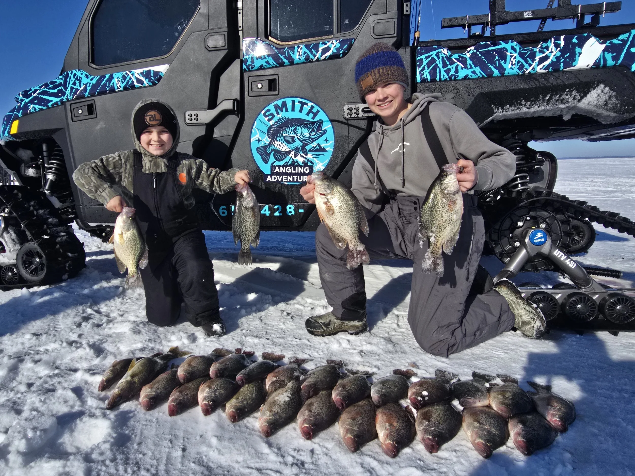Two people, a boy and an older male, standing on snow with a snowmobile behind them, displaying a catch of fish — a total of twelve fish with some lying on the snow and others held in their hands, in front of a lake or sea.