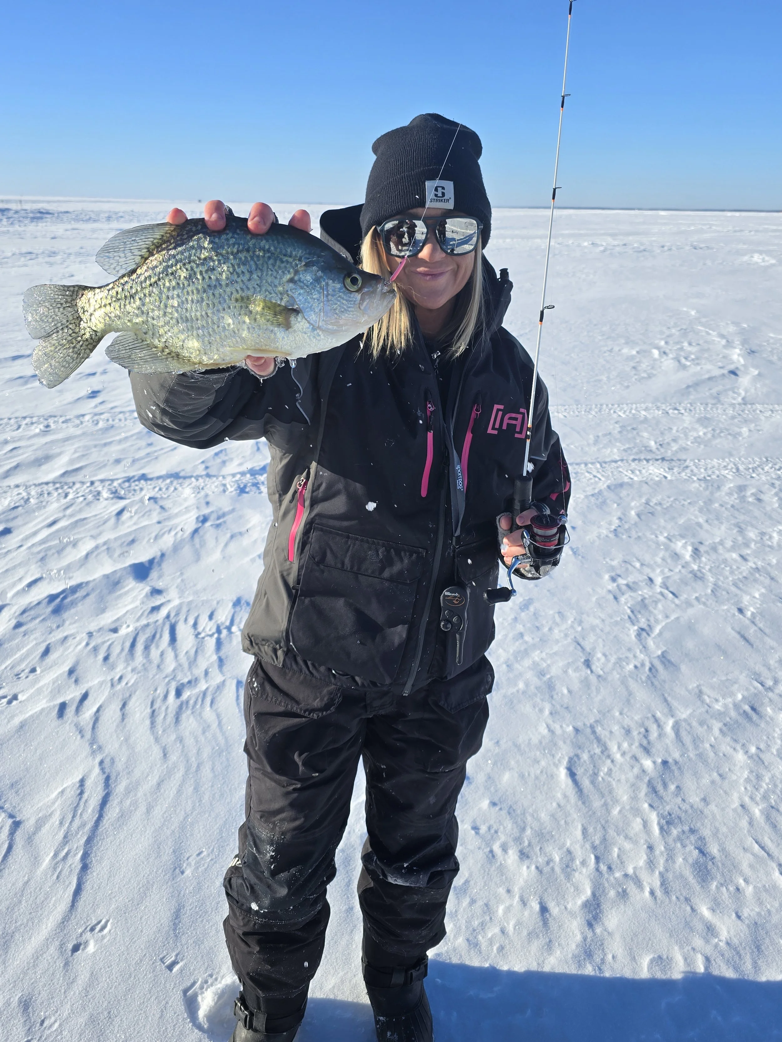 Woman dressed in winter clothing holding up a fish she caught on a snowy landscape in bright sunlight.