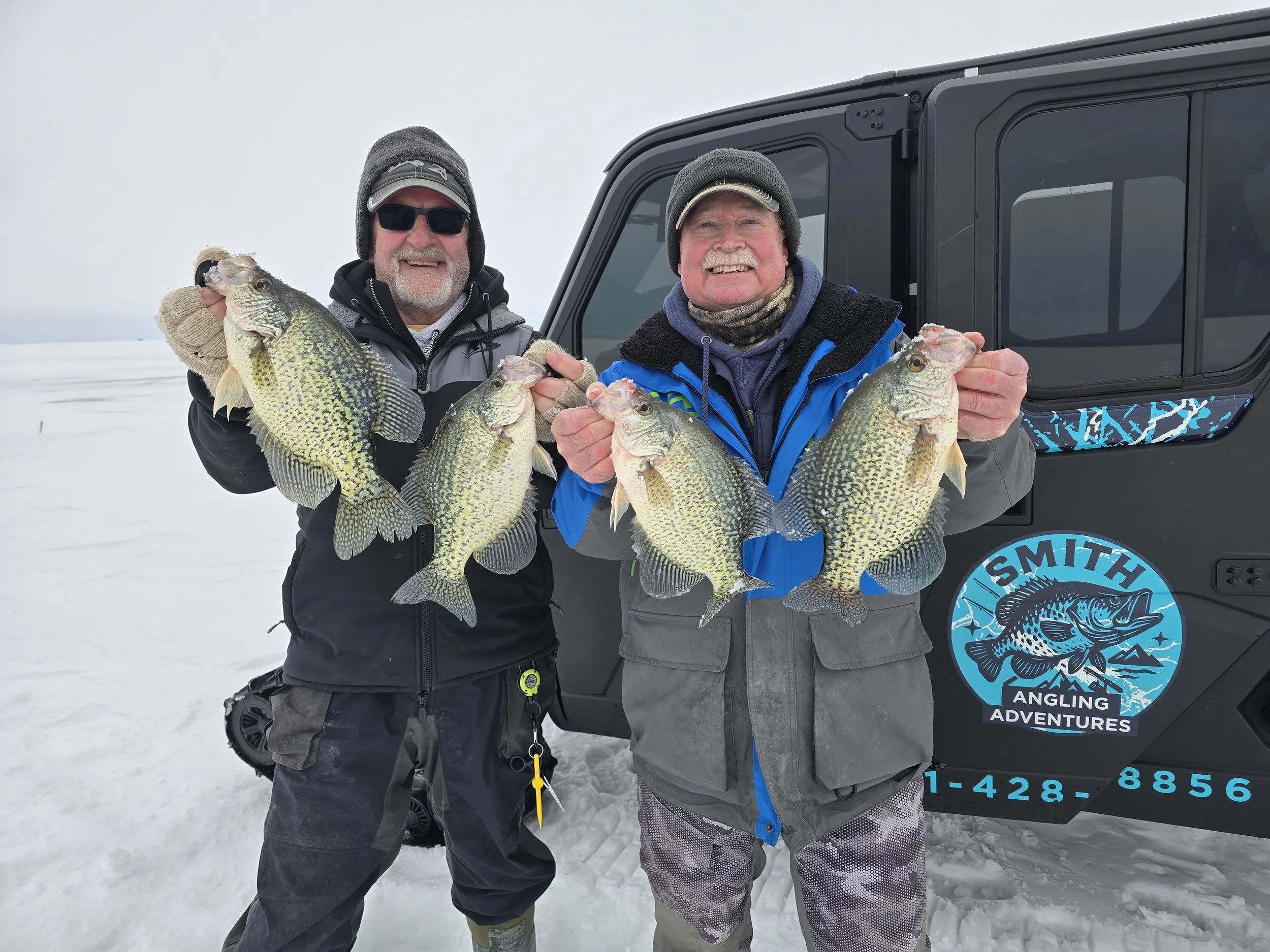 Two men stand on snow holding several large fish they caught. One man wears sunglasses and a beanie, the other wears a hat and a scarf. They are standing next to a vehicle with a logo that reads "Smith Angling Adventures".