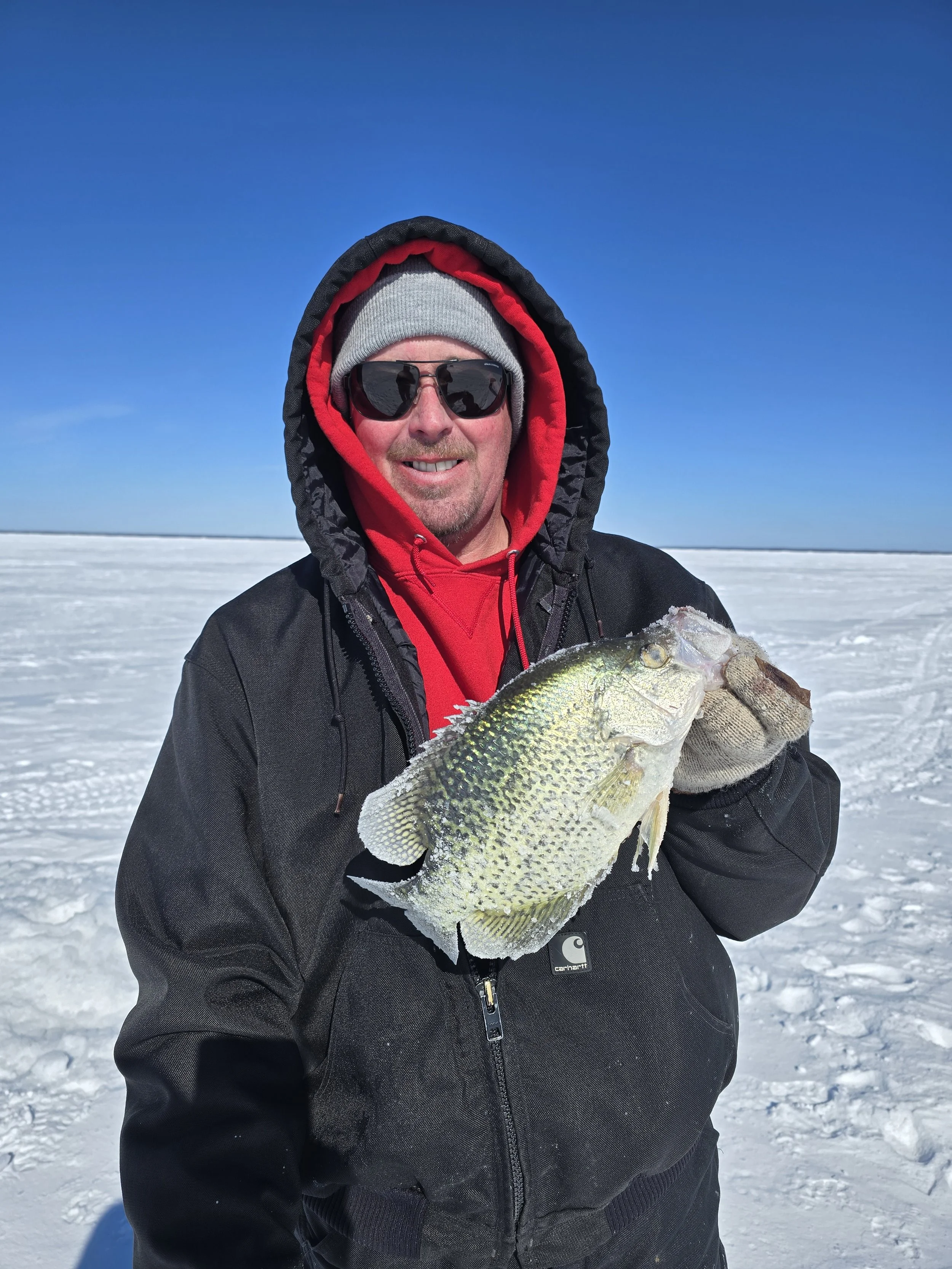 A man wearing sunglasses, a gray beanie, red hoodie, and black jacket holds a large fish on a snow-covered landscape with a clear blue sky in the background.