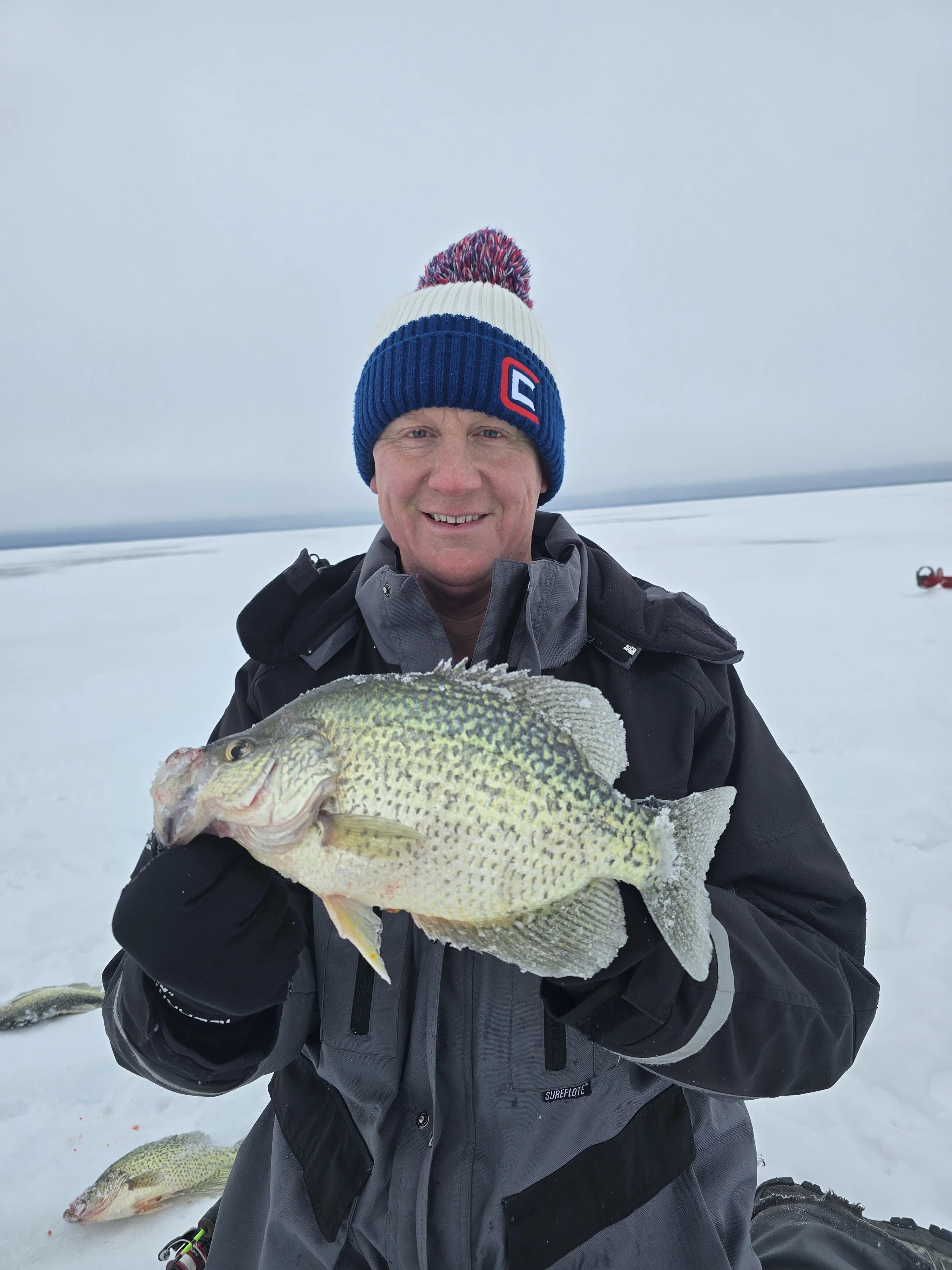 A man in winter clothing holding a large fish, standing on snow with a snowy, foggy background.