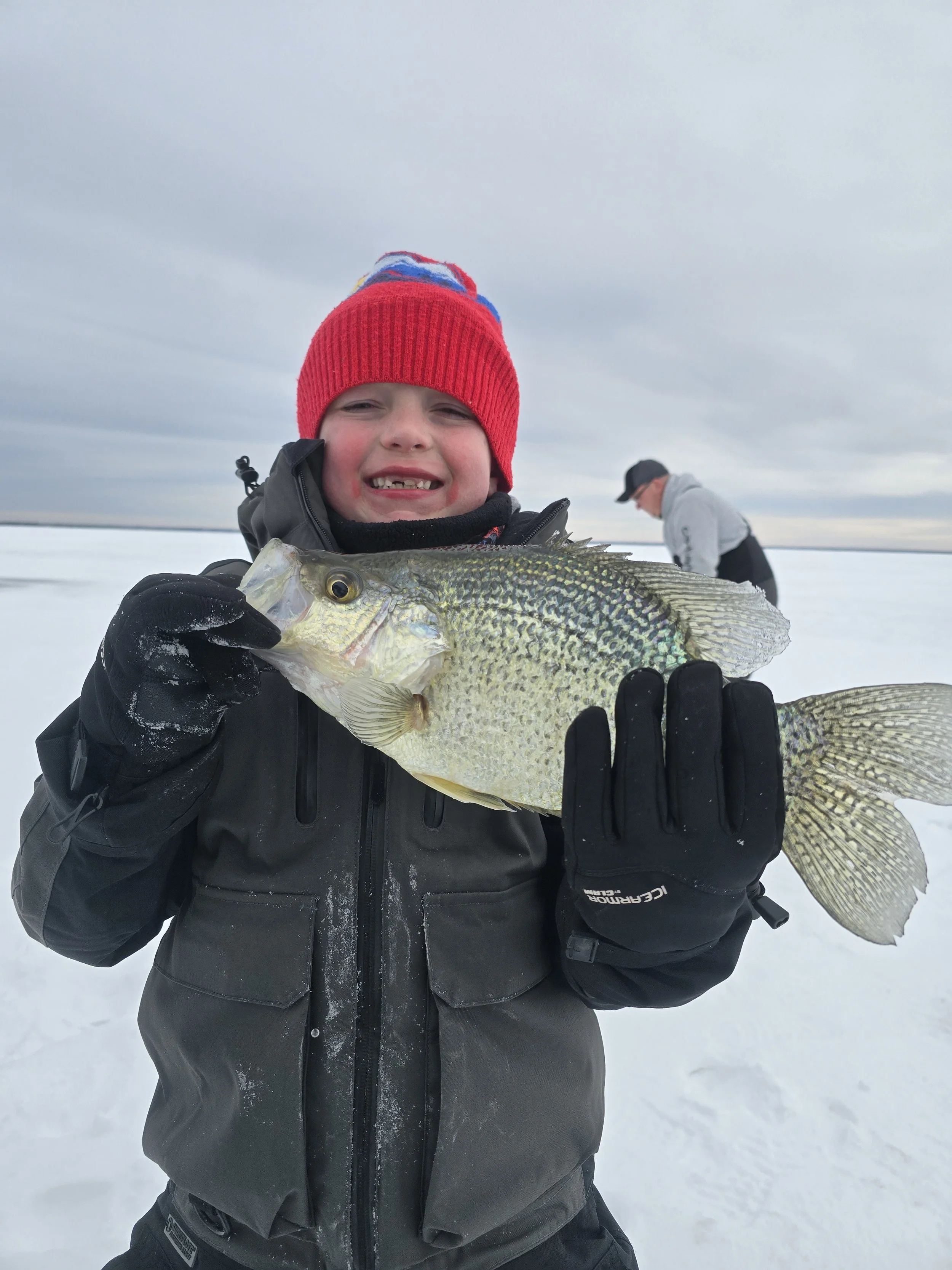 Young boy on a snowy ice-covered landscape, wearing a red winter hat with blue and red details, holding a large fish, smiling with missing front teeth, dressed in black winter gear, with a person in gray in the background.