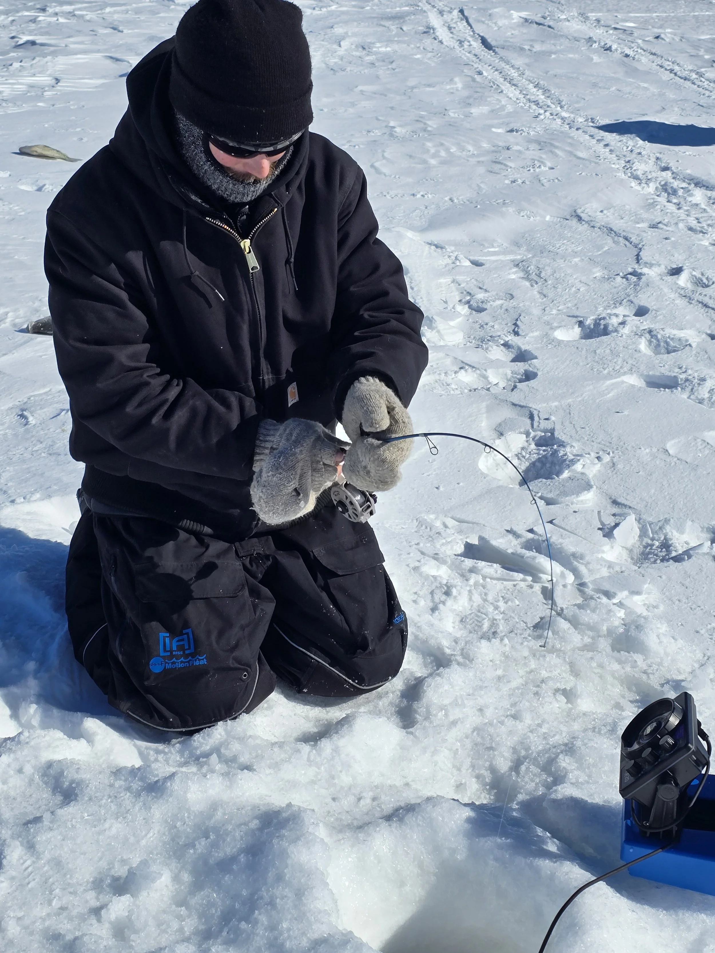 Man kneeling on snow, wearing black winter clothing, gloves, and a beanie, handling fishing equipment during winter.