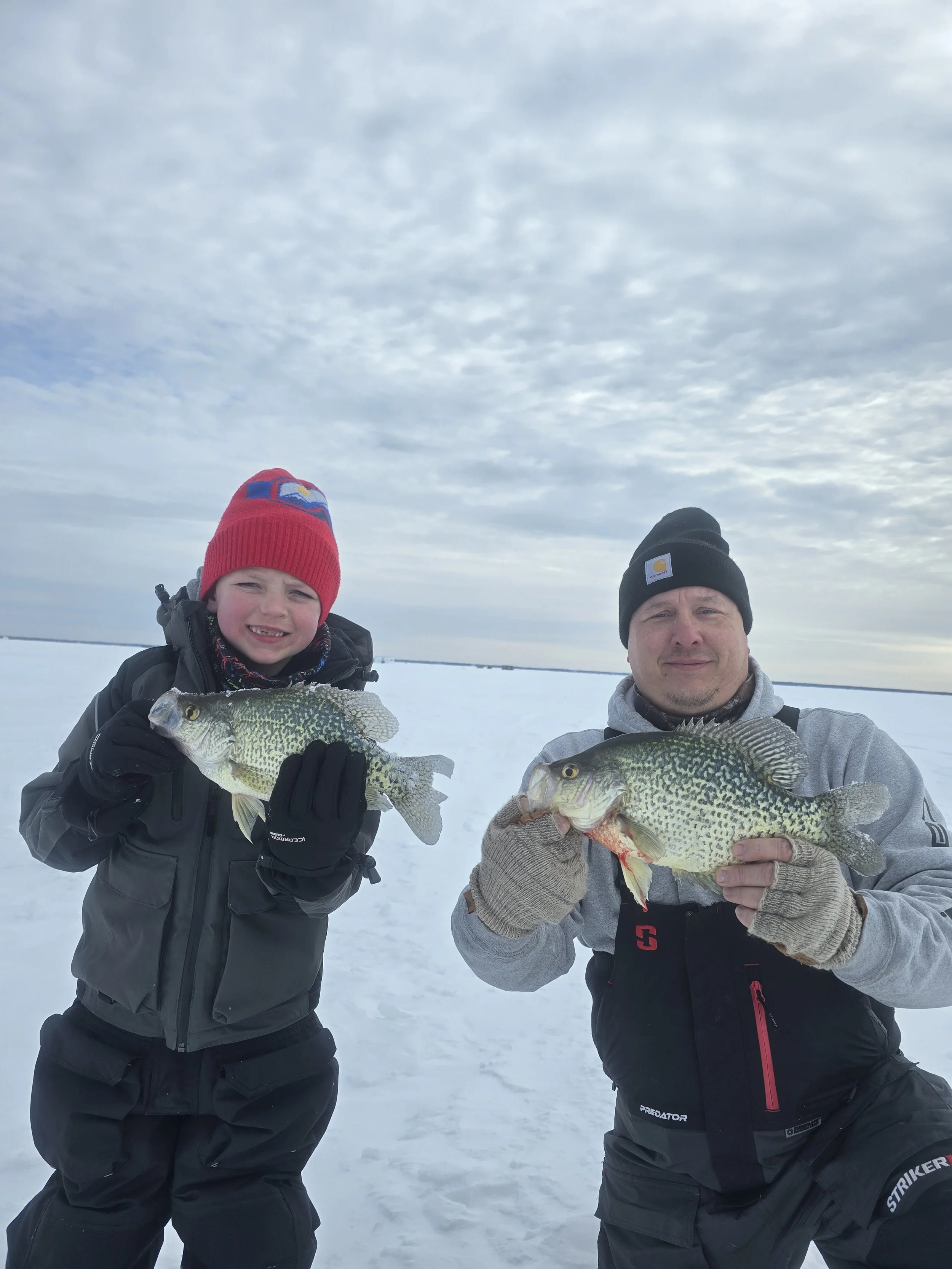A boy and a man standing on snow, each holding a large fish, with winter clothing and hats, under a cloudy sky.