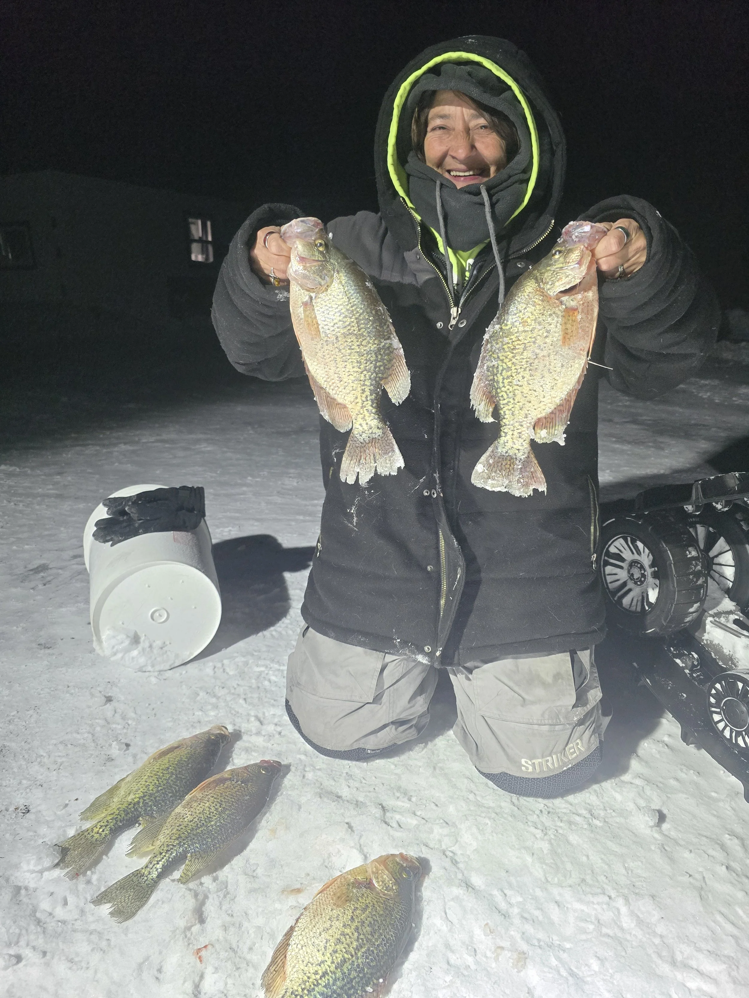 Person dressed in winter clothing holding two fish, with four more fish lying on snow-covered ground. A car and snowmobile are visible in the background at night.