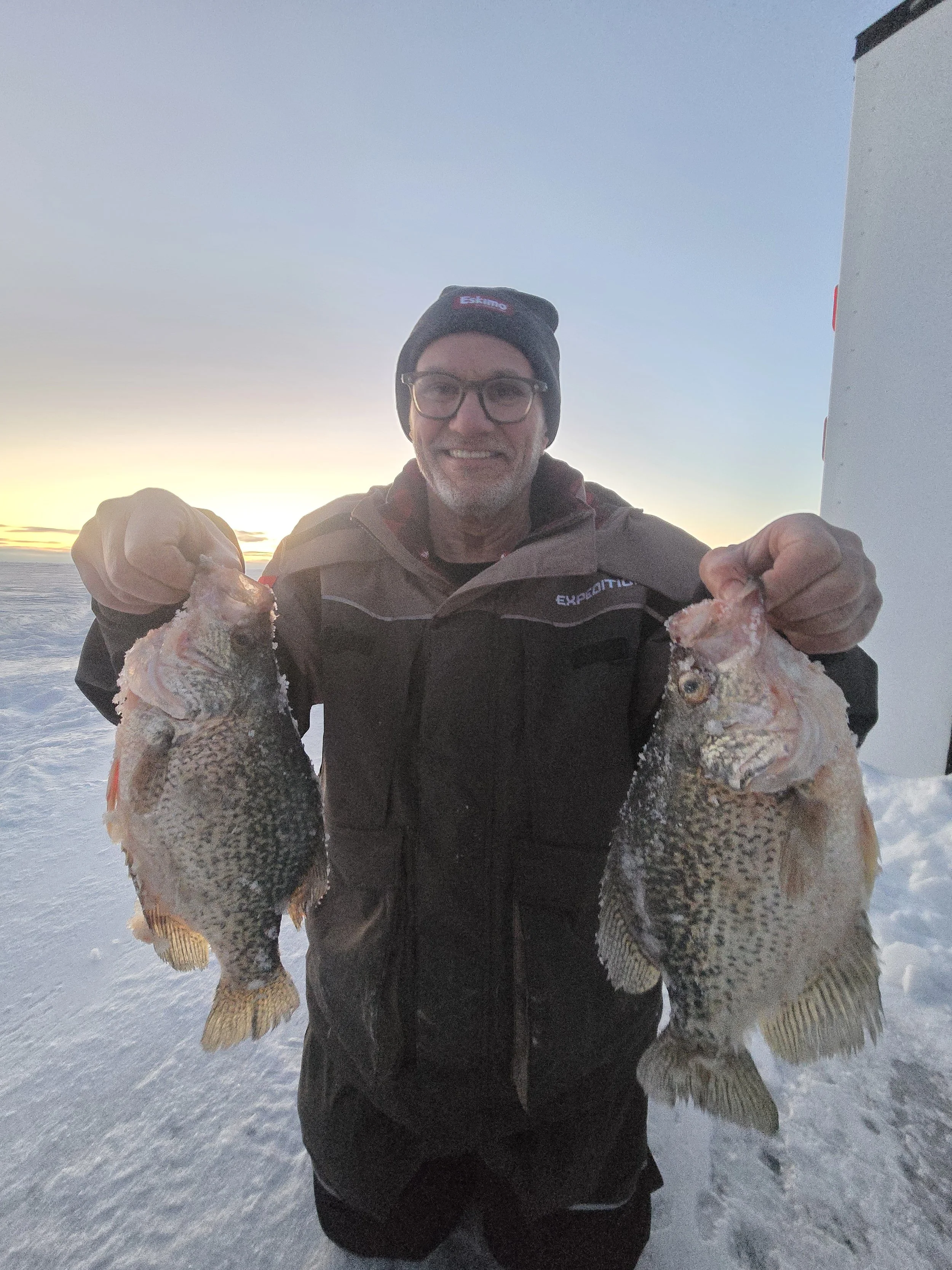 A person in winter clothing holding two large fish, standing on snow with a sunset and ocean in the background.