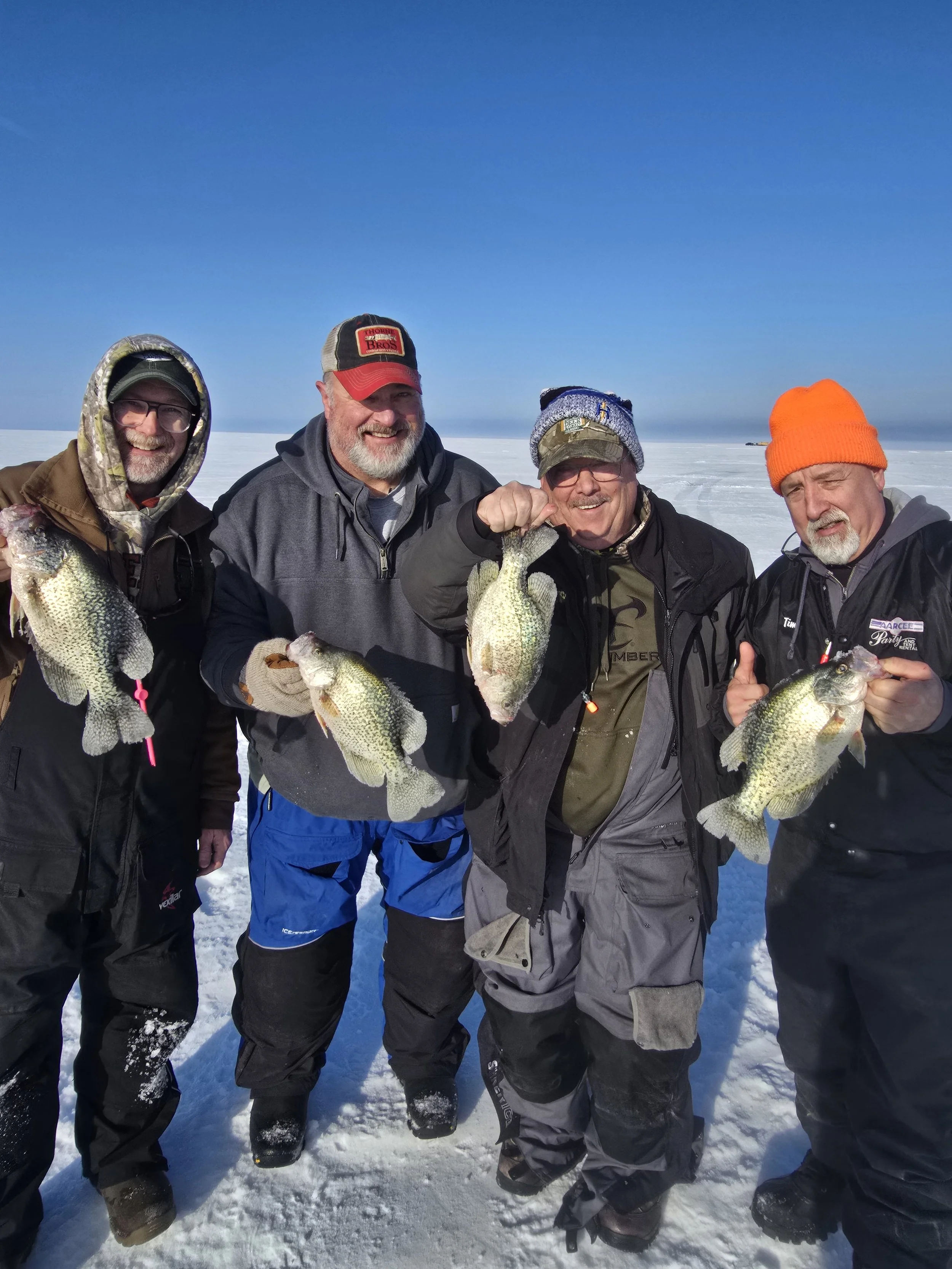Four men standing on a snowy landscape, holding large fish, dressed in winter clothing and smiling.