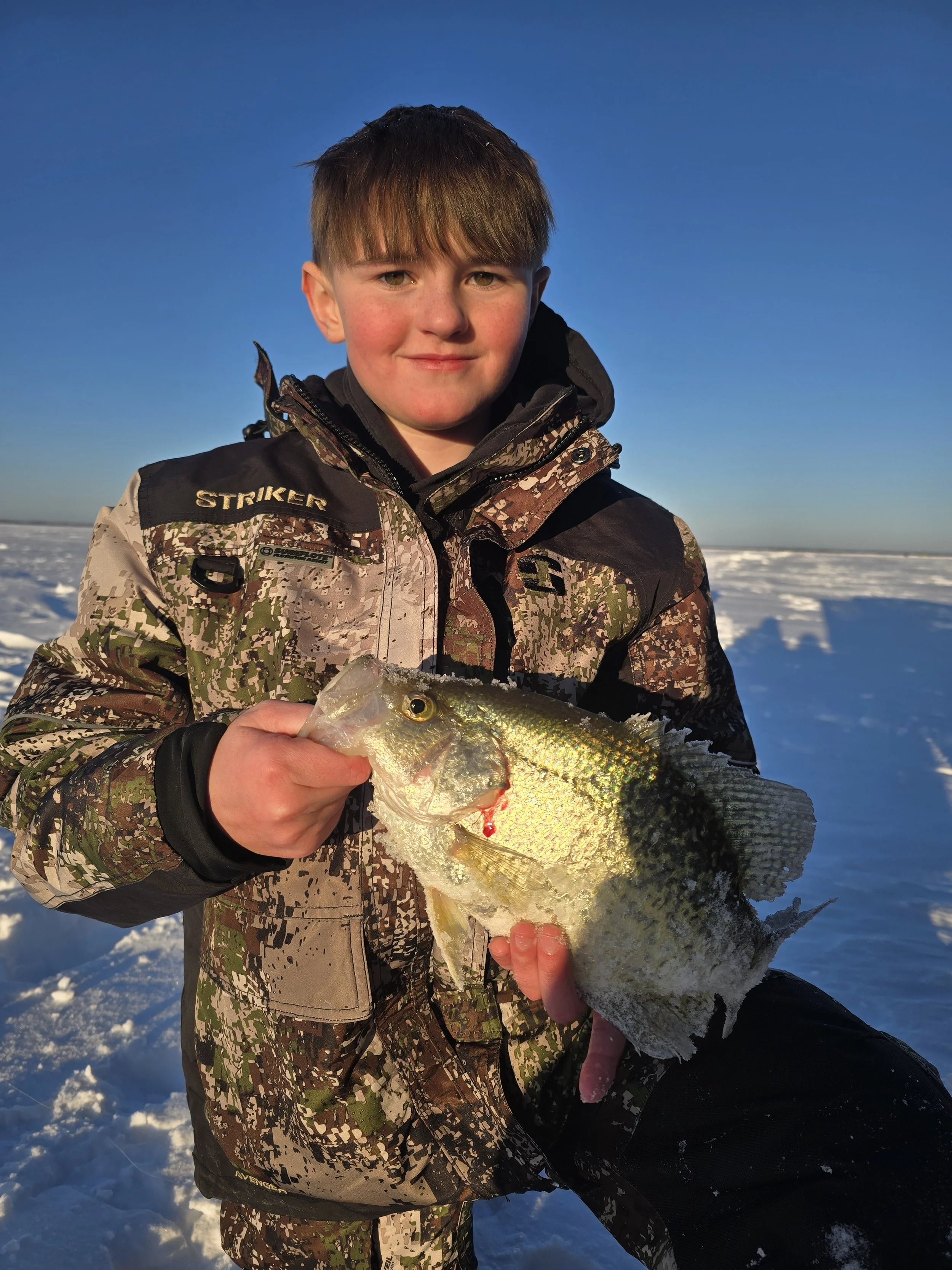 A young boy dressed in camouflage winter clothing holding a largemouth bass fish outdoors on a snowy landscape with a clear blue sky.
