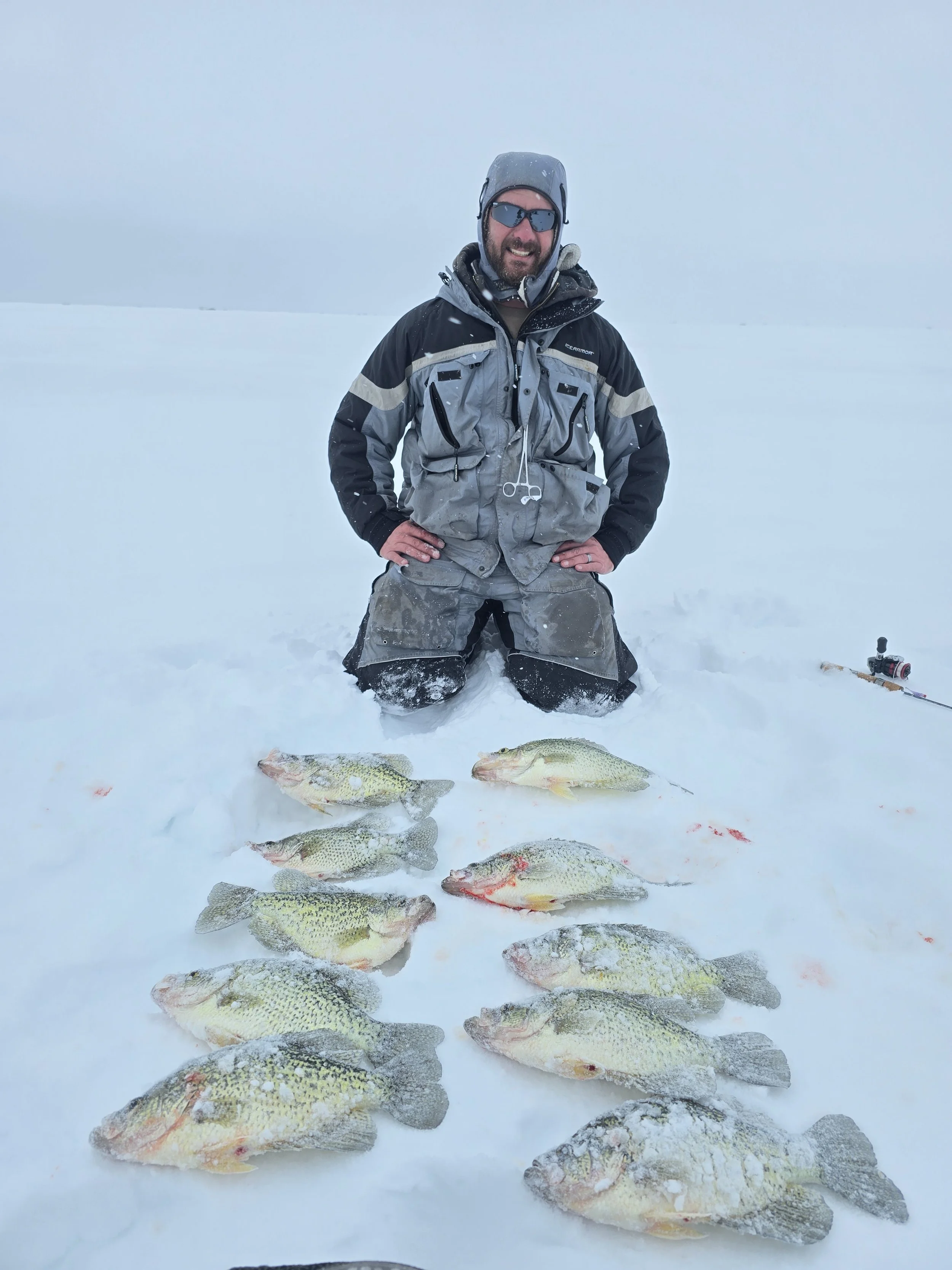 A man kneeling in snow wearing winter gear and sunglasses, smiling, with ten fish laid out in front of him on the snow. The fish are likely caught from ice fishing.