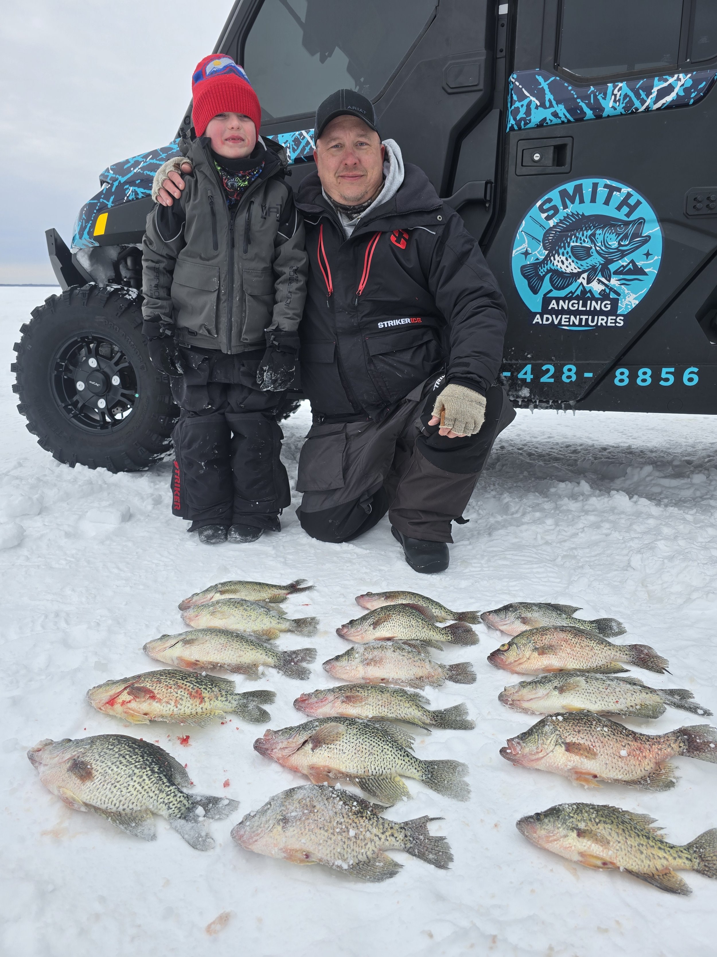 Two people, an adult man and a young boy, kneeling on snow next to a line of caught fish, with a black vehicle marked with a blue and white logo for Smith Angling Adventures in the background.