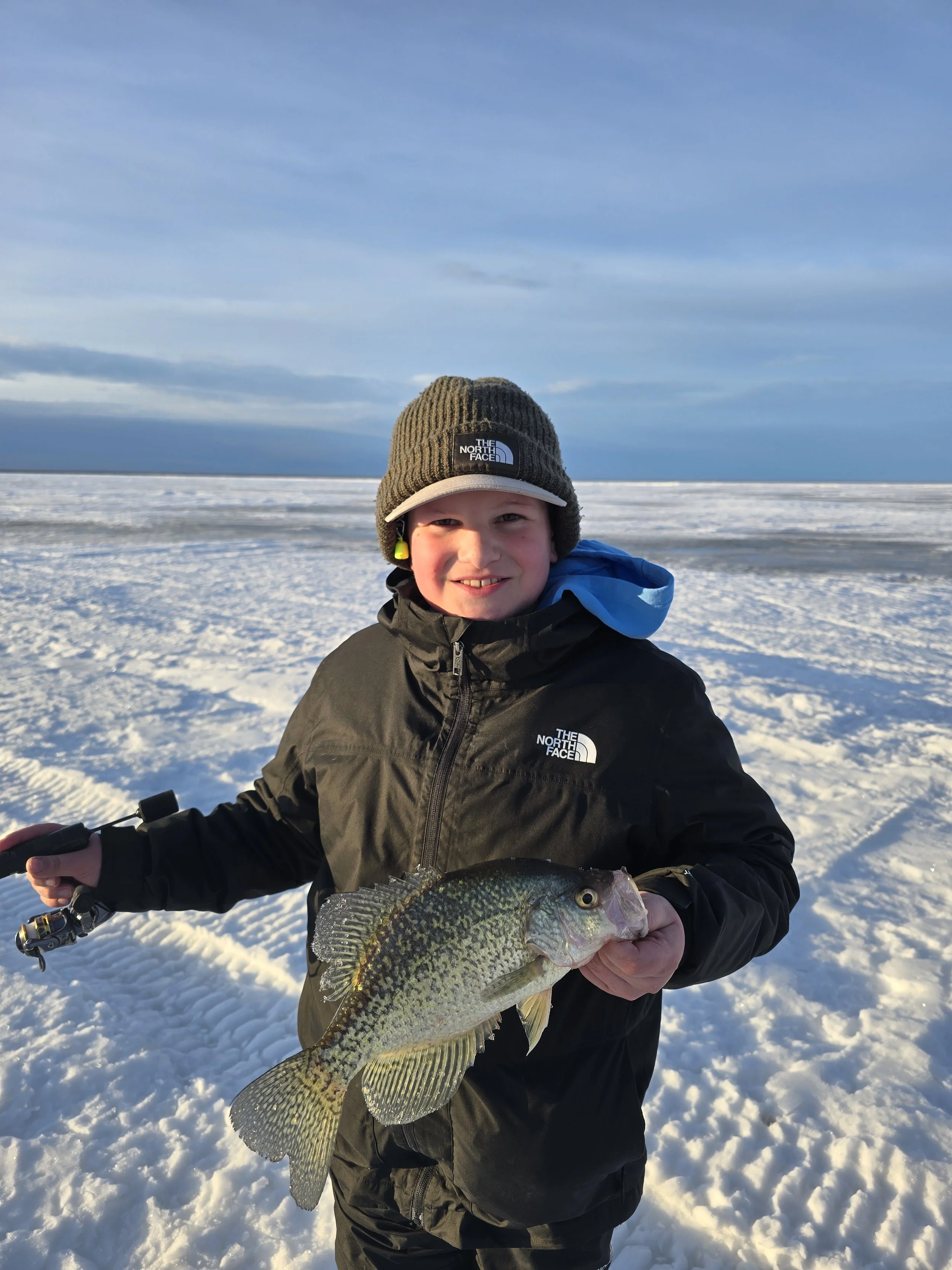 A young boy standing on a snowy landscape holding a large fish, wearing a black North Face winter jacket and a brown knitted hat, smiling at the camera, with a partly cloudy sky in the background.