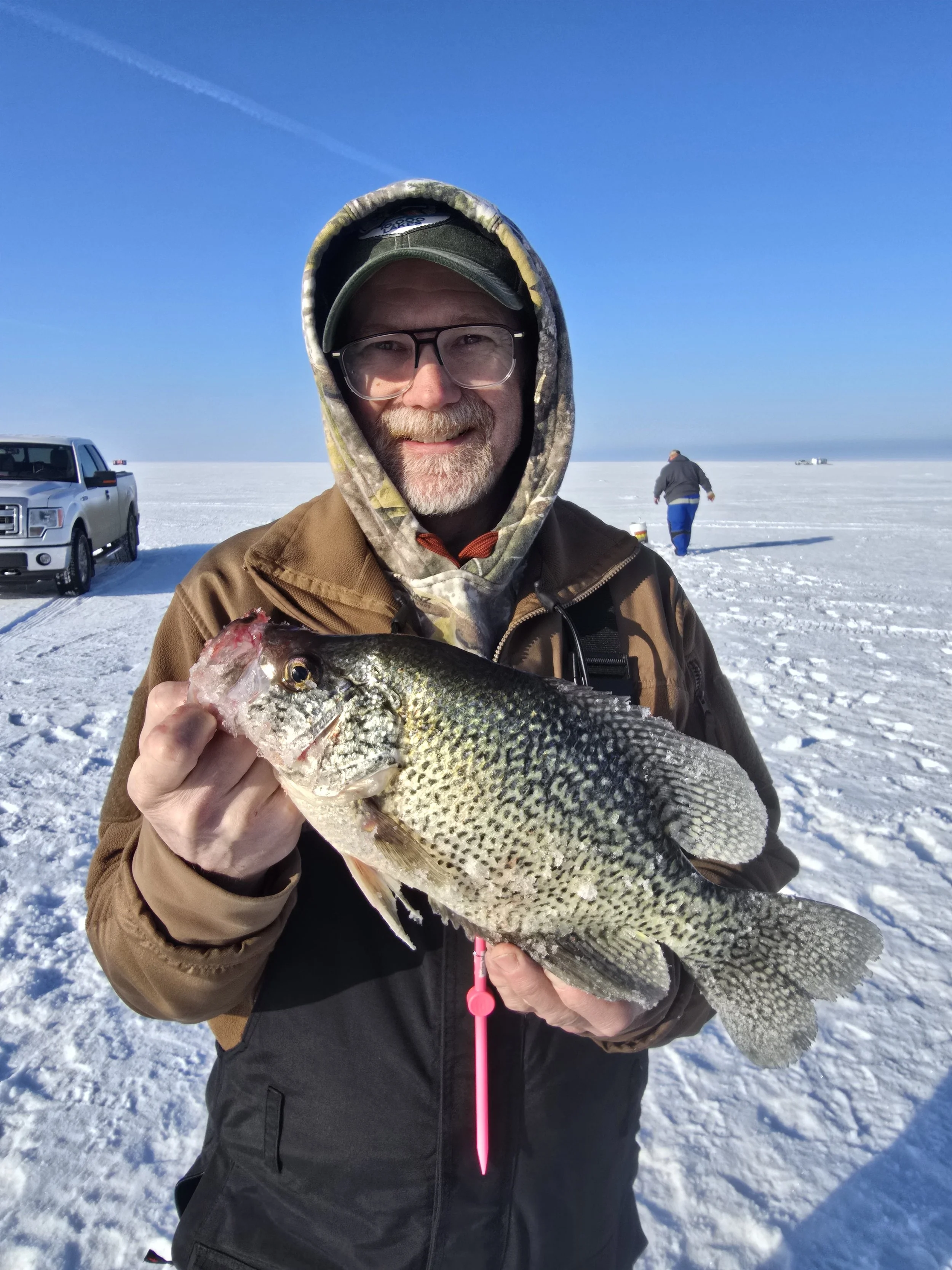 Man in winter clothing holding a large fish on a snowy landscape with clear blue sky, a vehicle, and another person in the background.