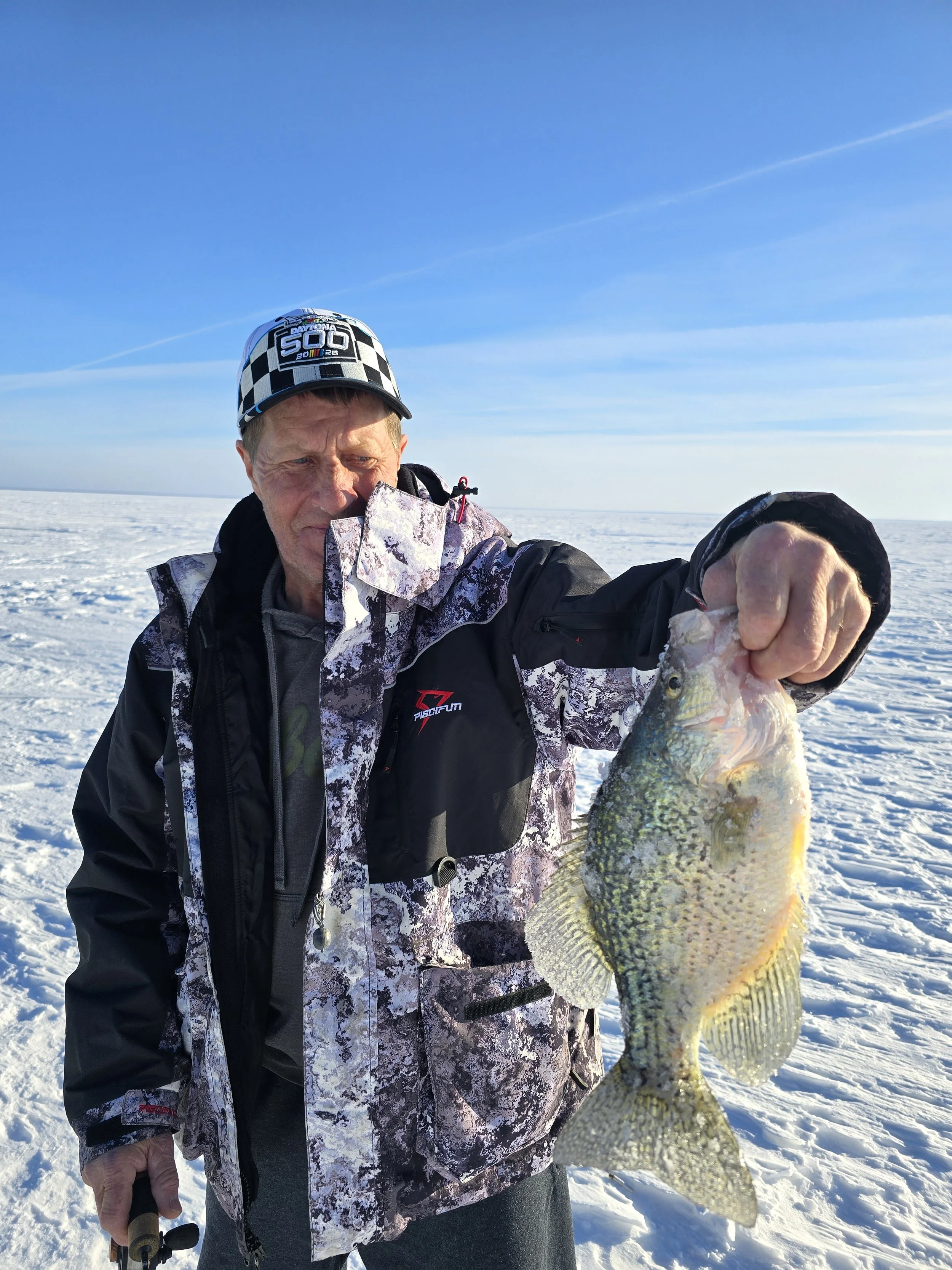 A man wearing a checkered cap and camouflage jacket holding a large fish on a snowy landscape under a clear blue sky.