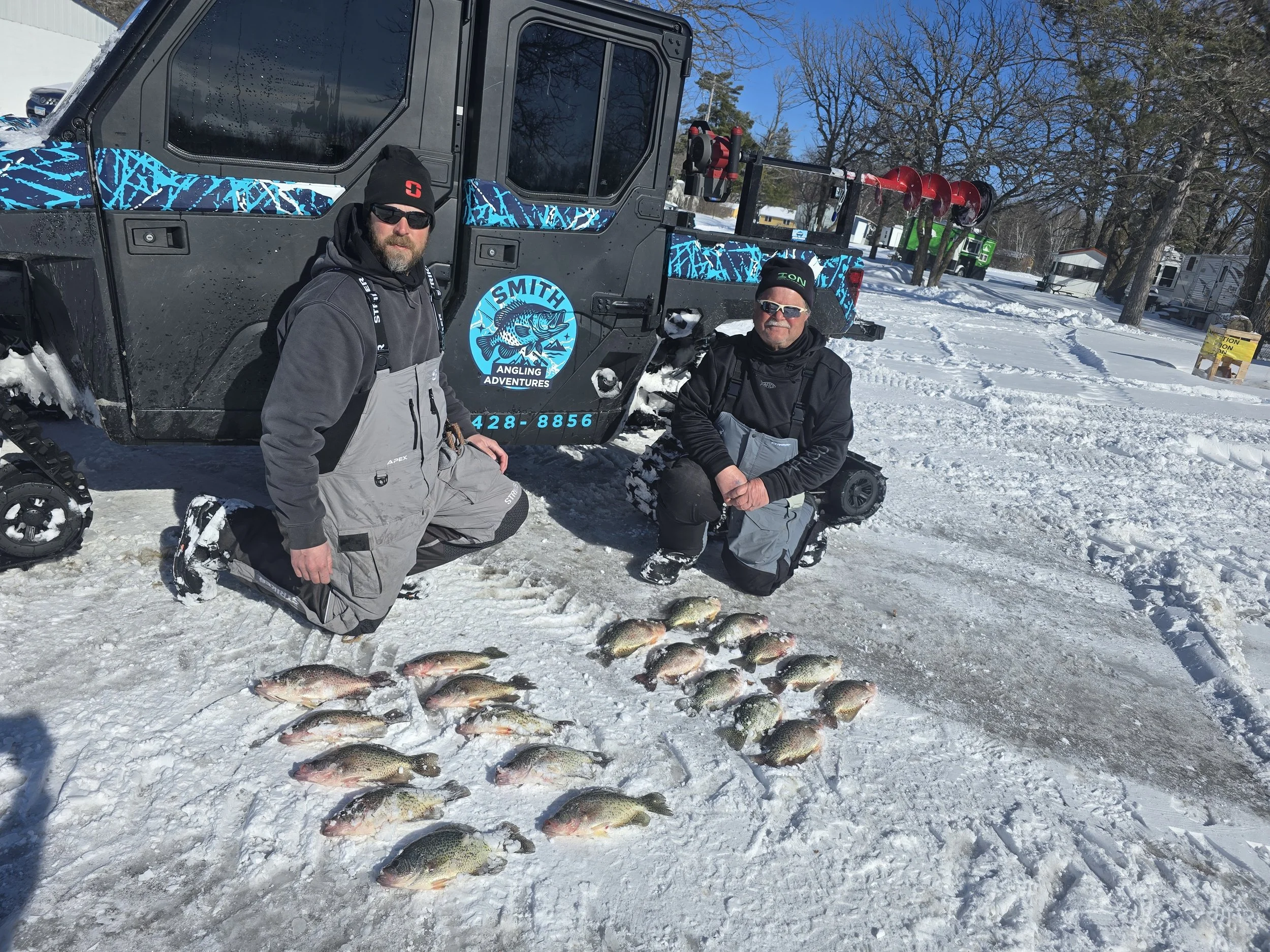 Two men in winter fishing gear kneeling in snow in front of a large catch of fish on the ice, with an ice fishing vehicle and trees in the background.