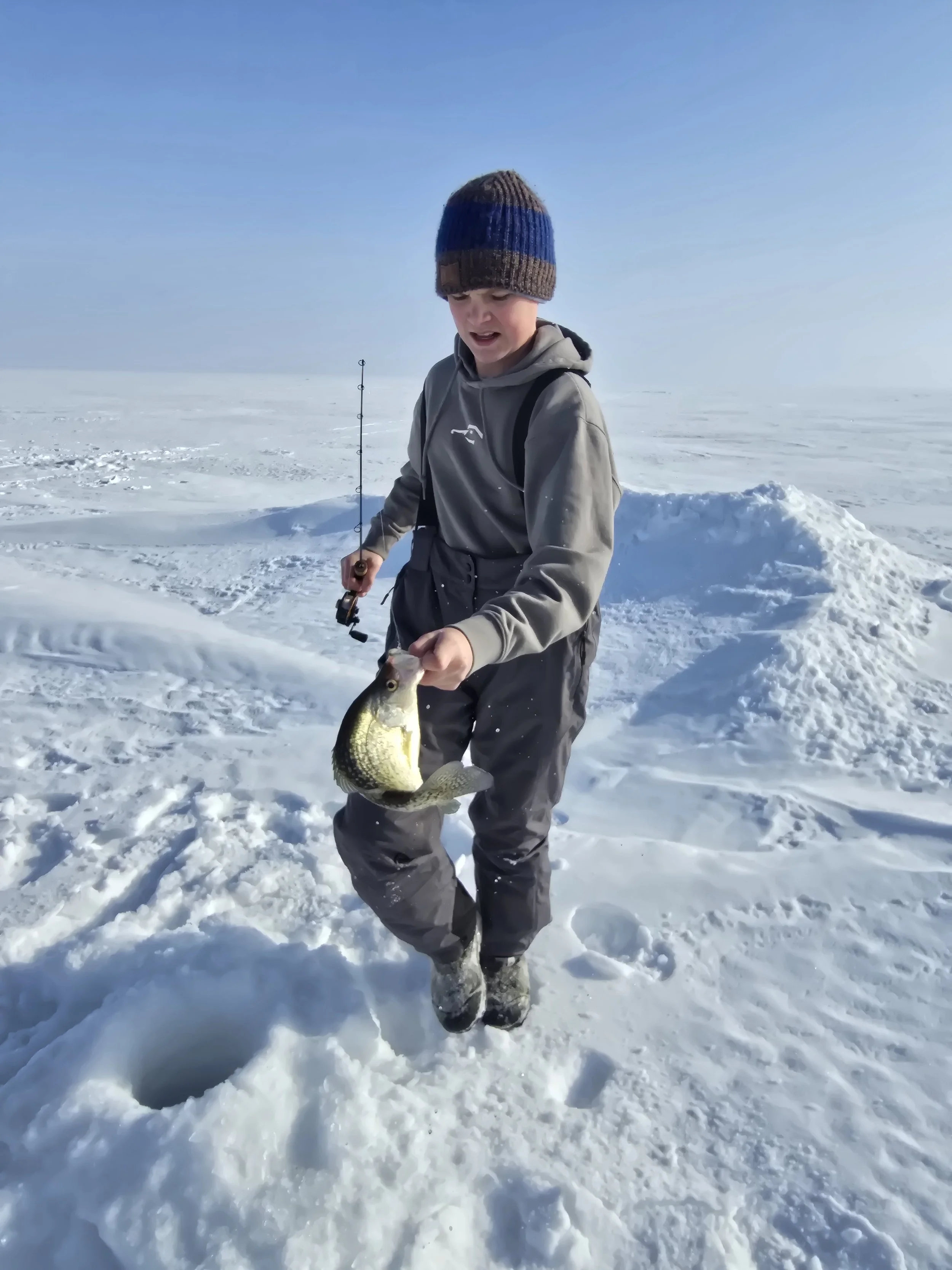 A young boy dressed in winter clothing holding a fish he caught in a snowy outdoor landscape.