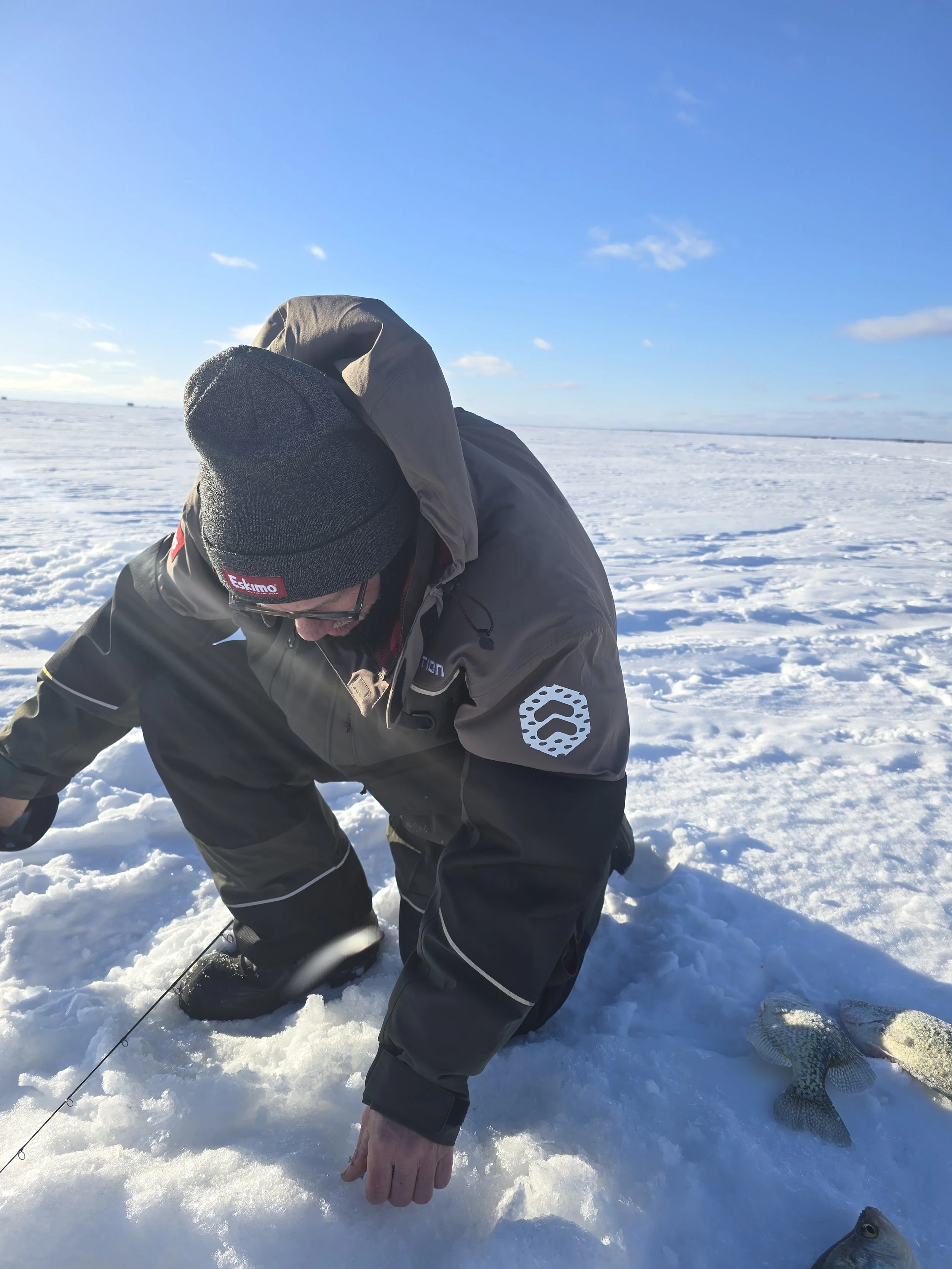 A person dressed in winter clothing, including a beanie and a jacket, is kneeling on snow and holding a fishing line, with a fish lying on the snow nearby. The background shows a vast snowy landscape under a clear blue sky.