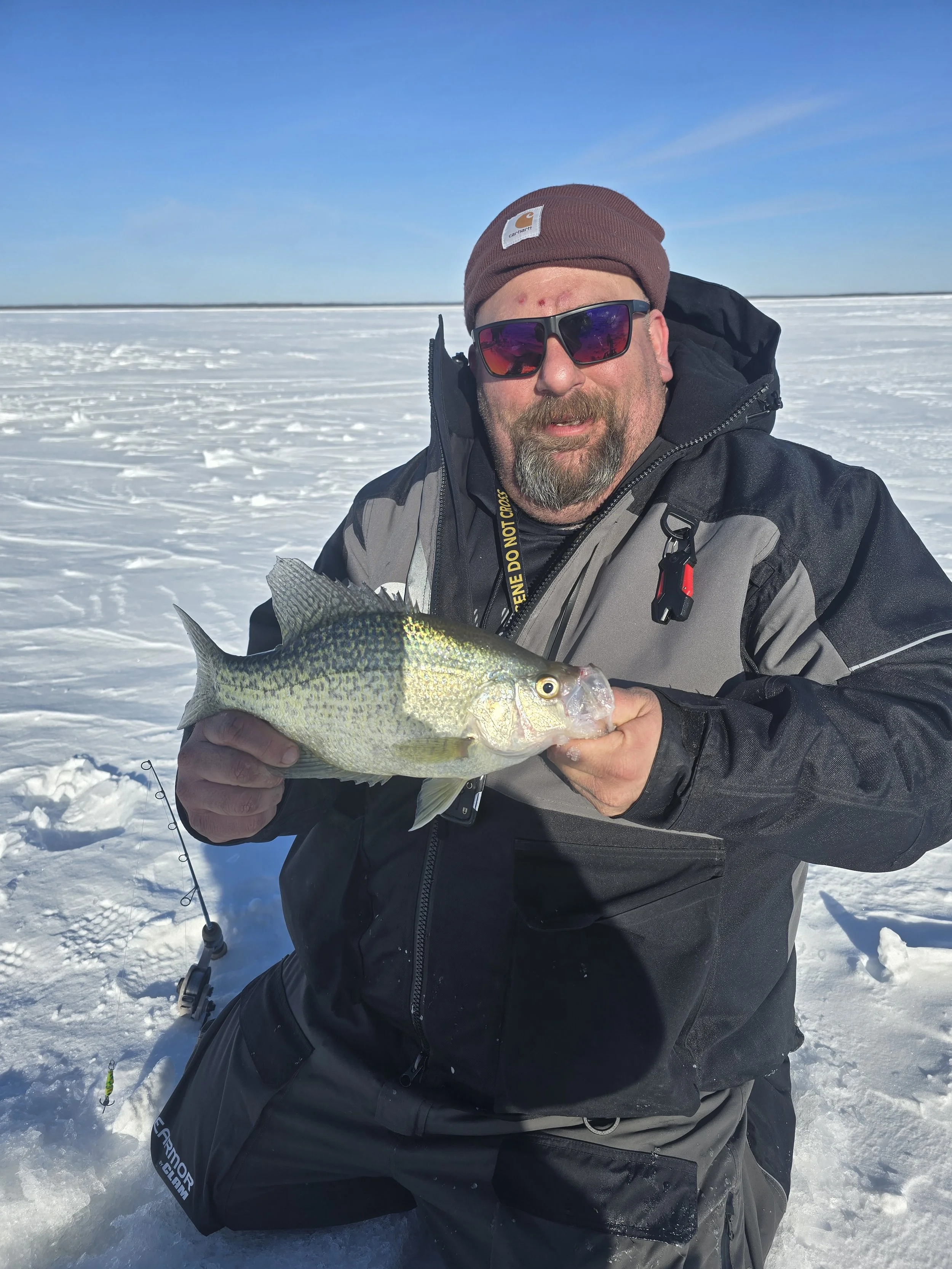 A man in winter gear holding a fish on a snowy landscape with a clear blue sky.