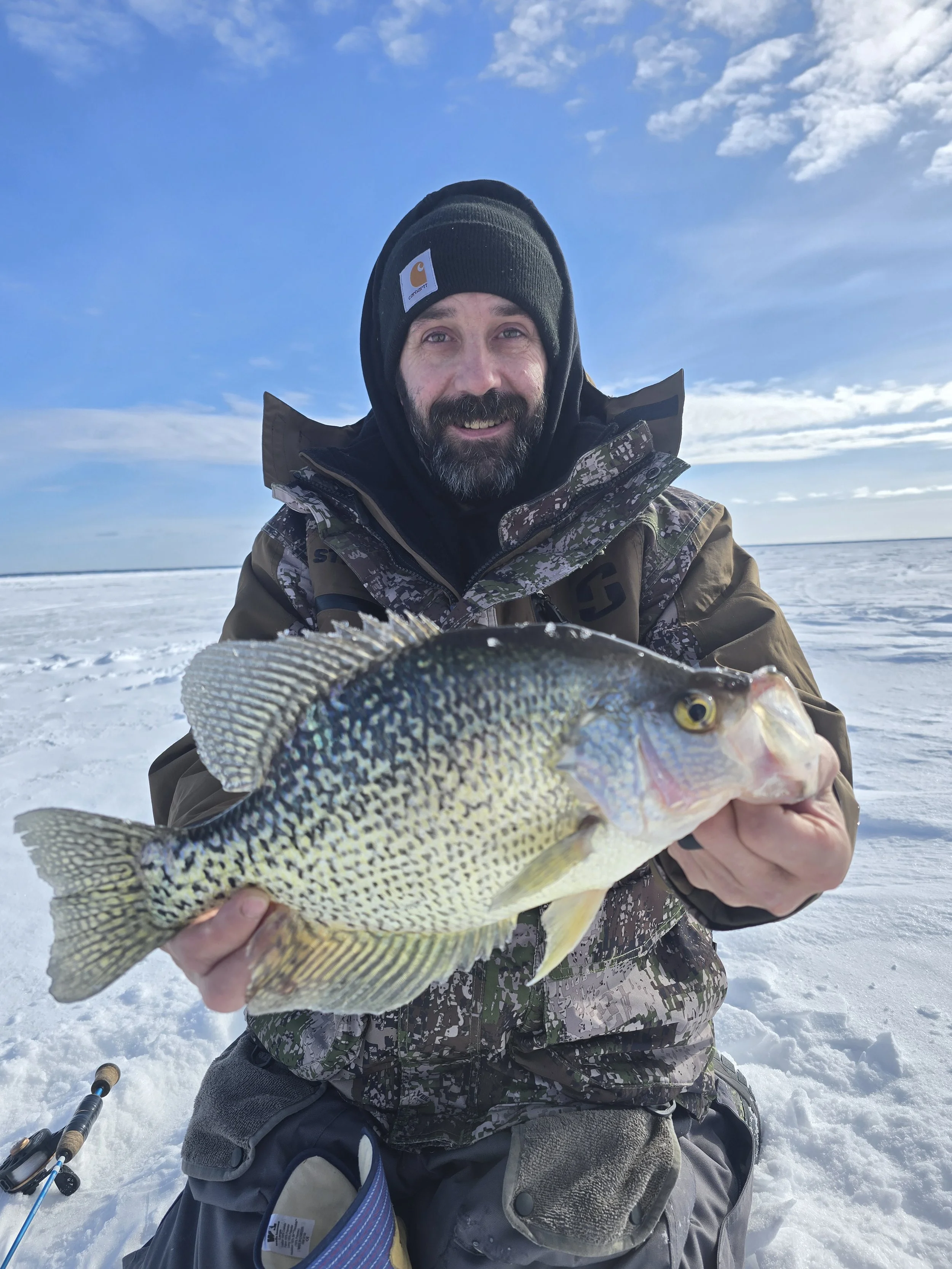 A man in winter clothing, including a Carhartt beanie and camouflage jacket, holding a fish in front of a snowy landscape with a clear blue sky and some clouds.