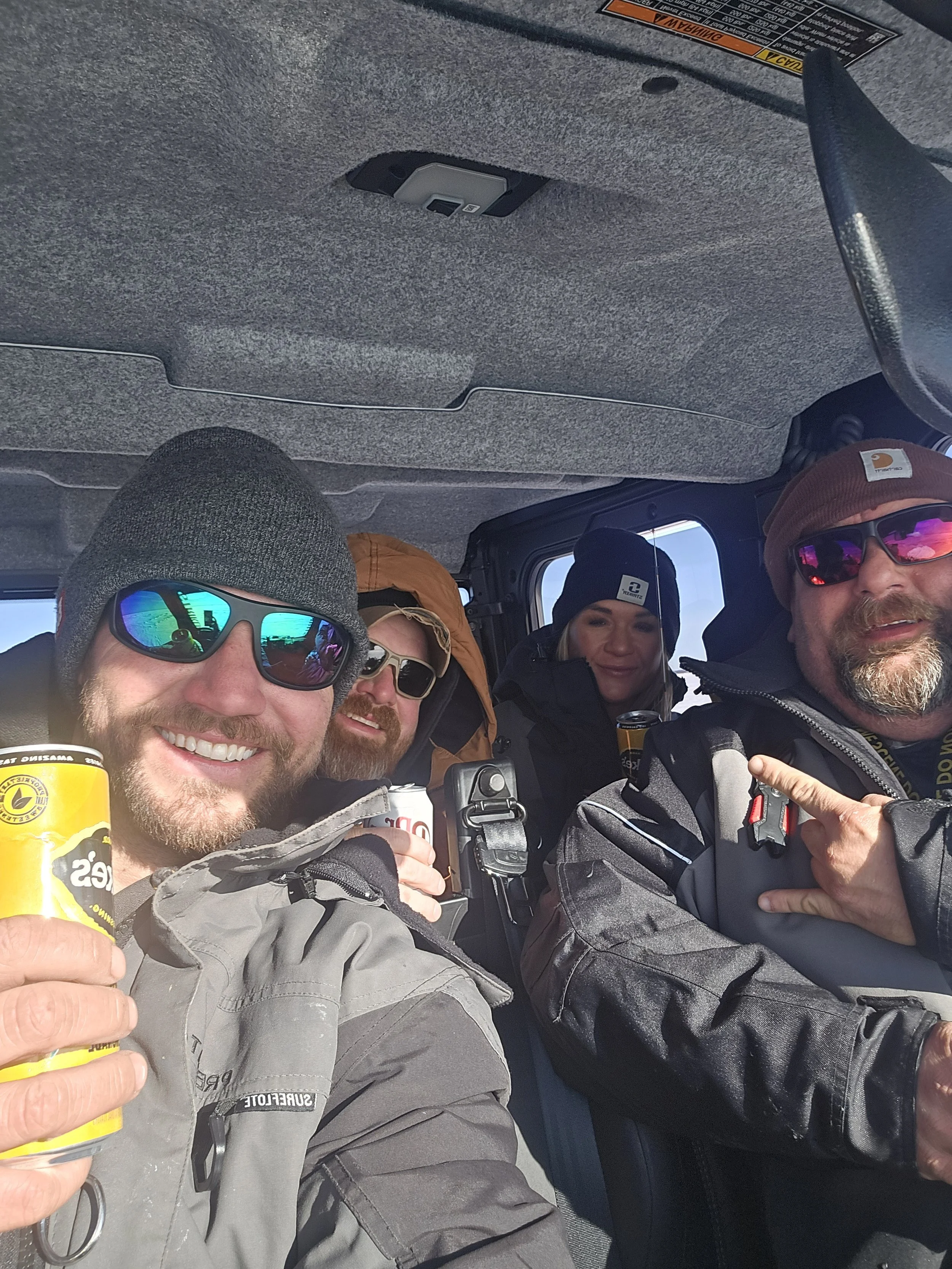 A group of four people inside a vehicle, smiling and holding drinks, wearing outdoor gear including jackets, hats, and sunglasses.
