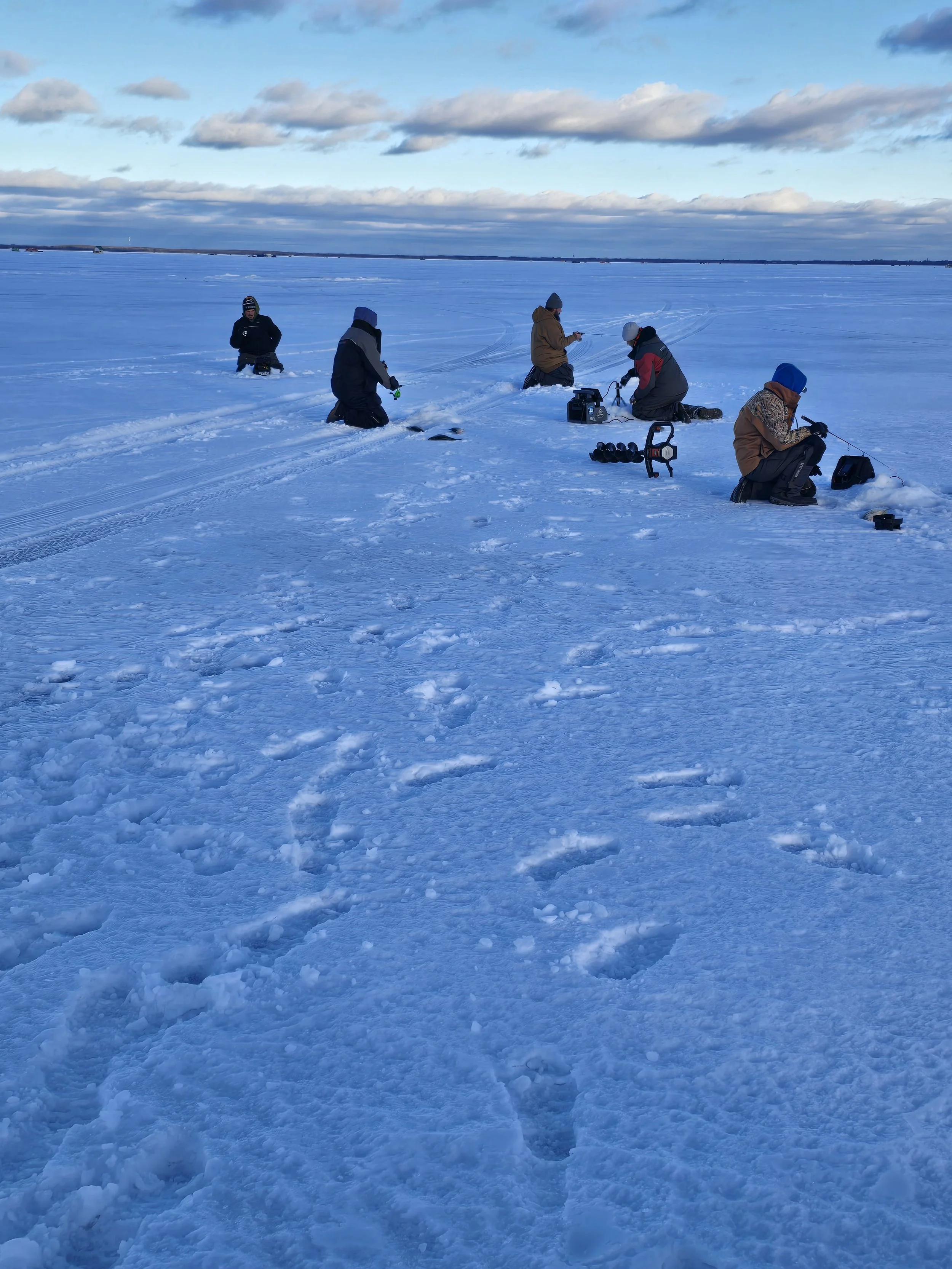 People ice fishing on a frozen lake with snow, wearing winter clothing, in scenic winter landscape under cloudy sky.