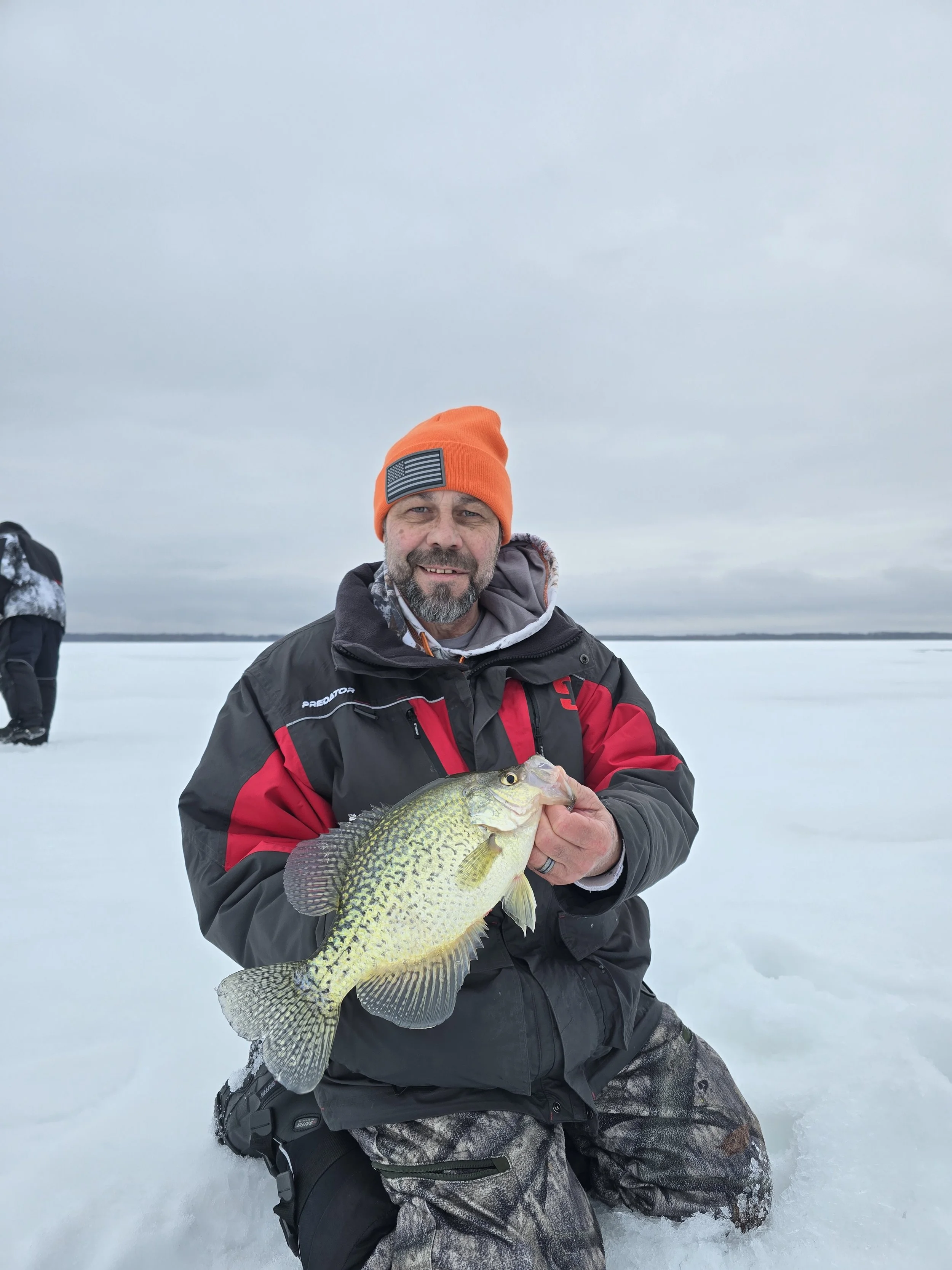 Man in winter clothing holding a fish on a snowy landscape under an overcast sky.