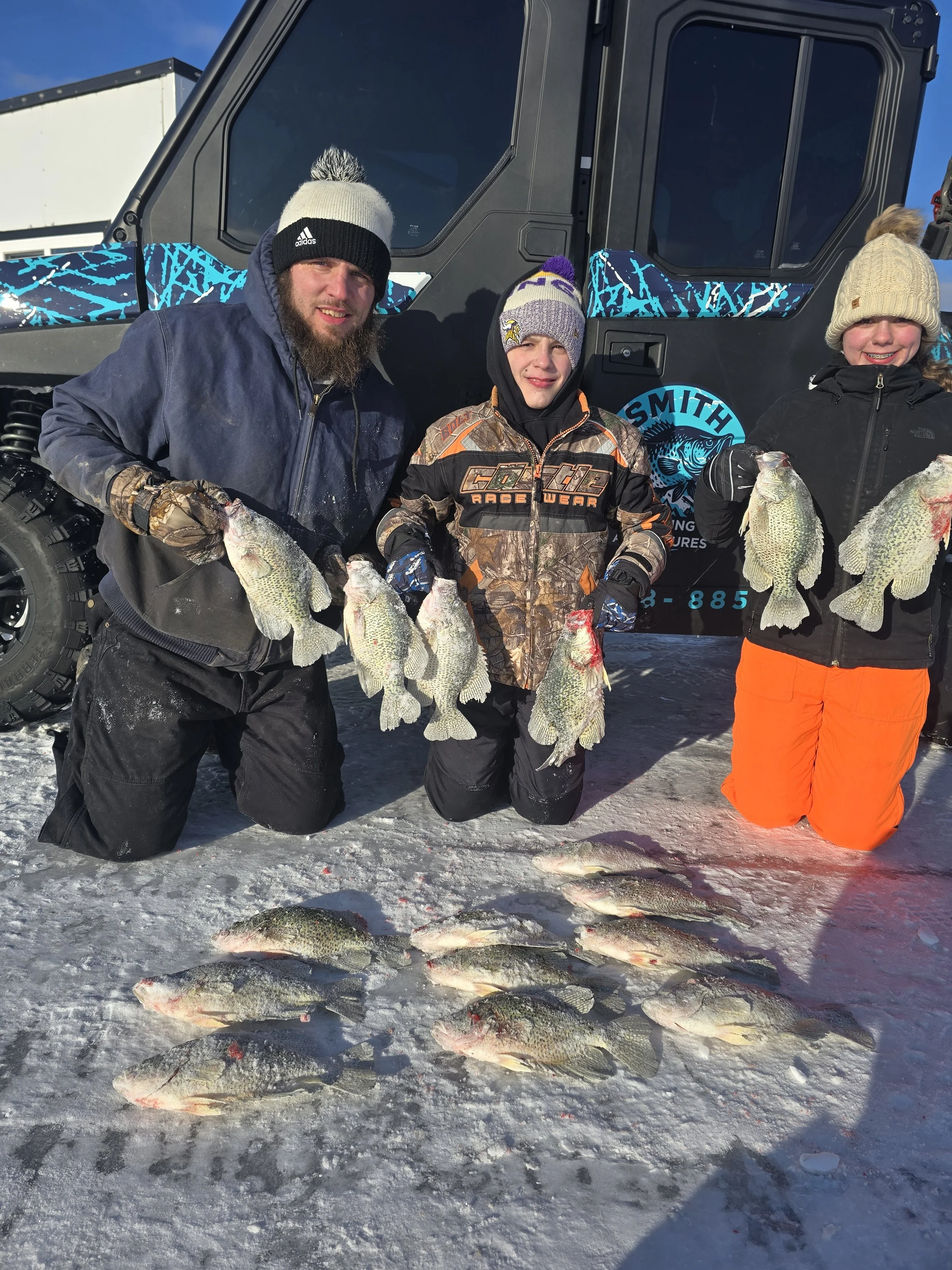 Three people kneeling on snow with fish they caught, in winter clothing, in front of an off-road vehicle outdoors.