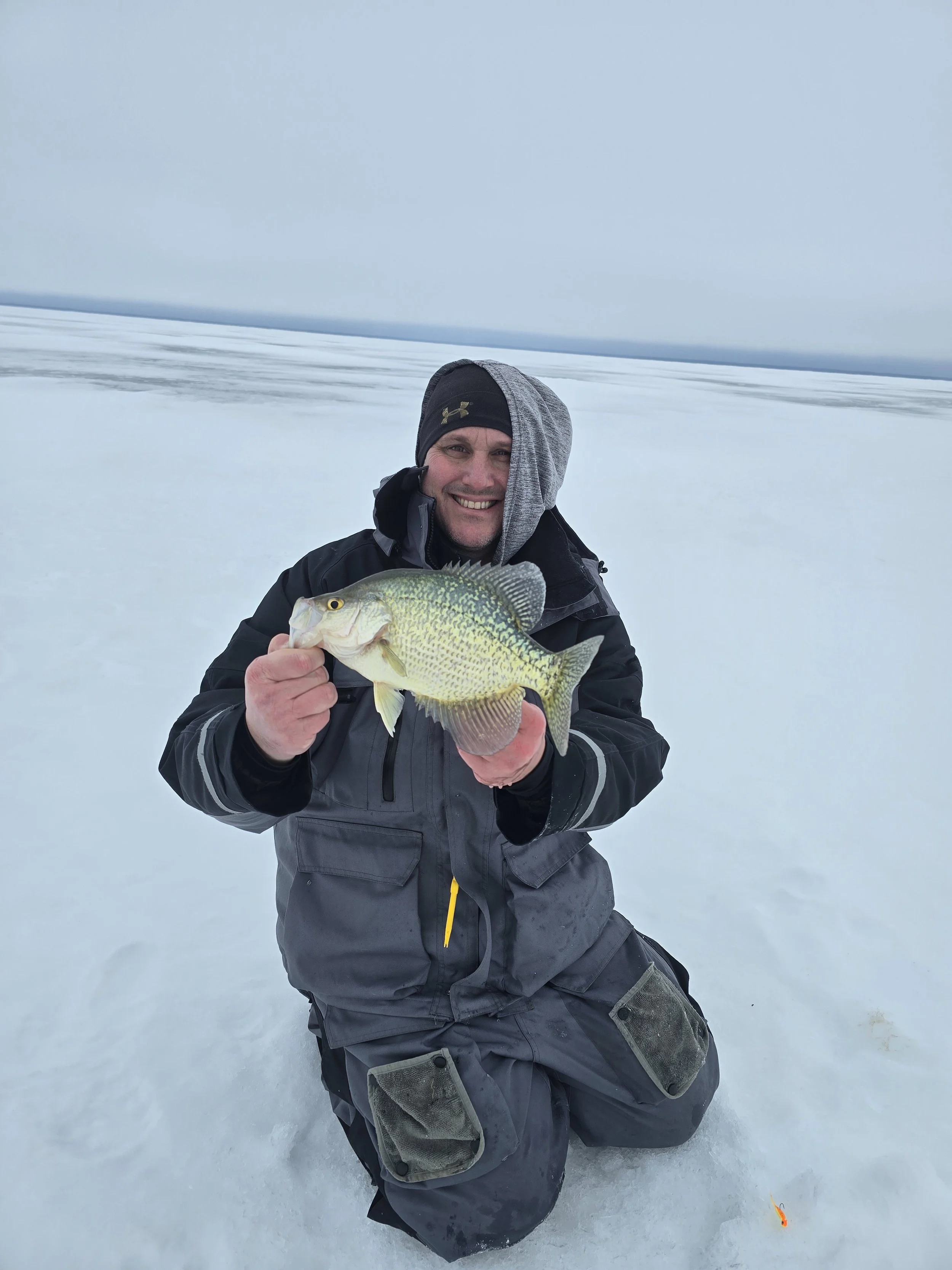 A man kneeling on snowy ice holding a fish he caught, with a cloudy sky and icy landscape in the background.