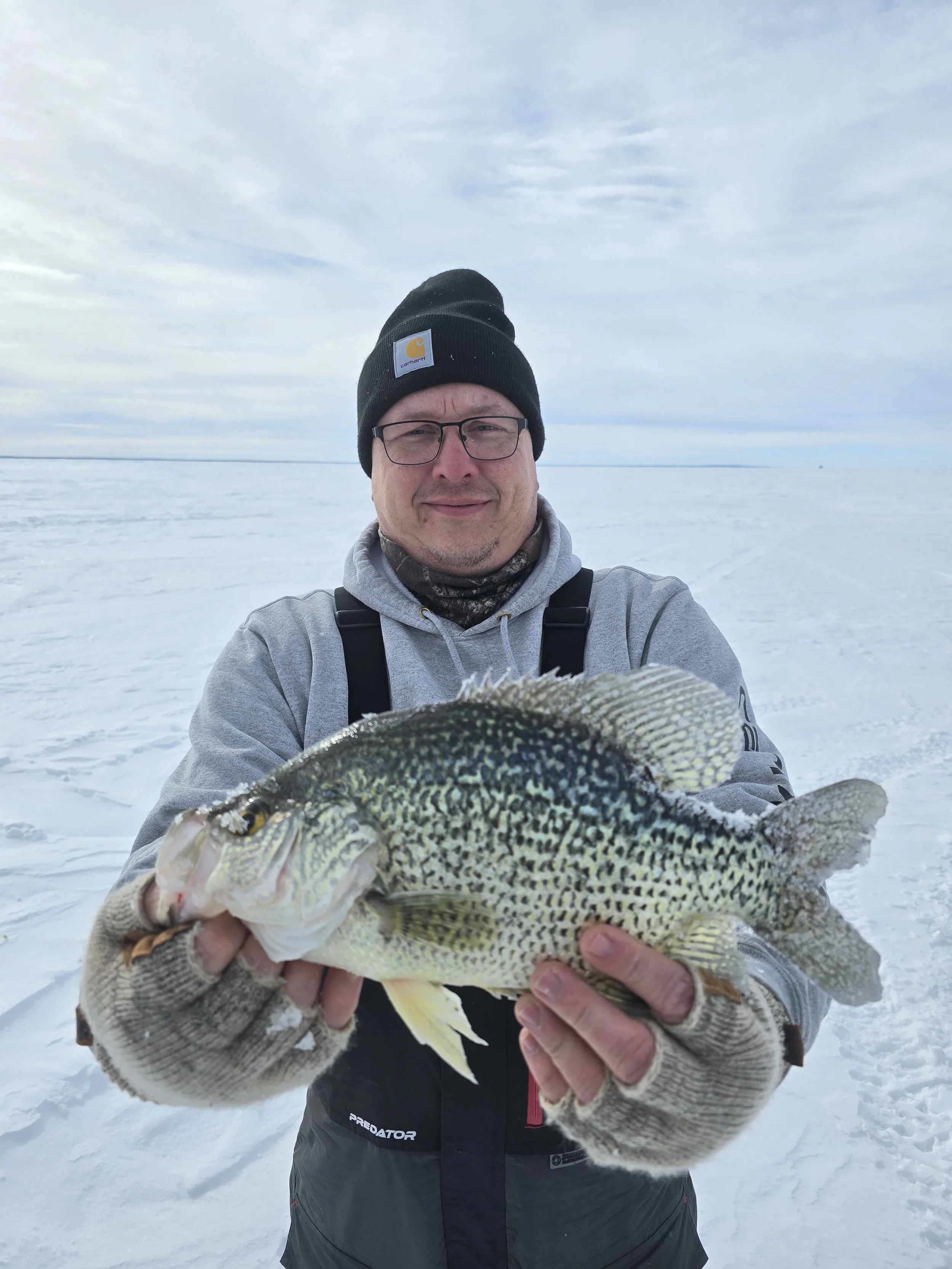 A man wearing a black Carhartt beanie, glasses, and a gray hoodie kneels on snow and holds a large fish with black, yellow, and white scales.