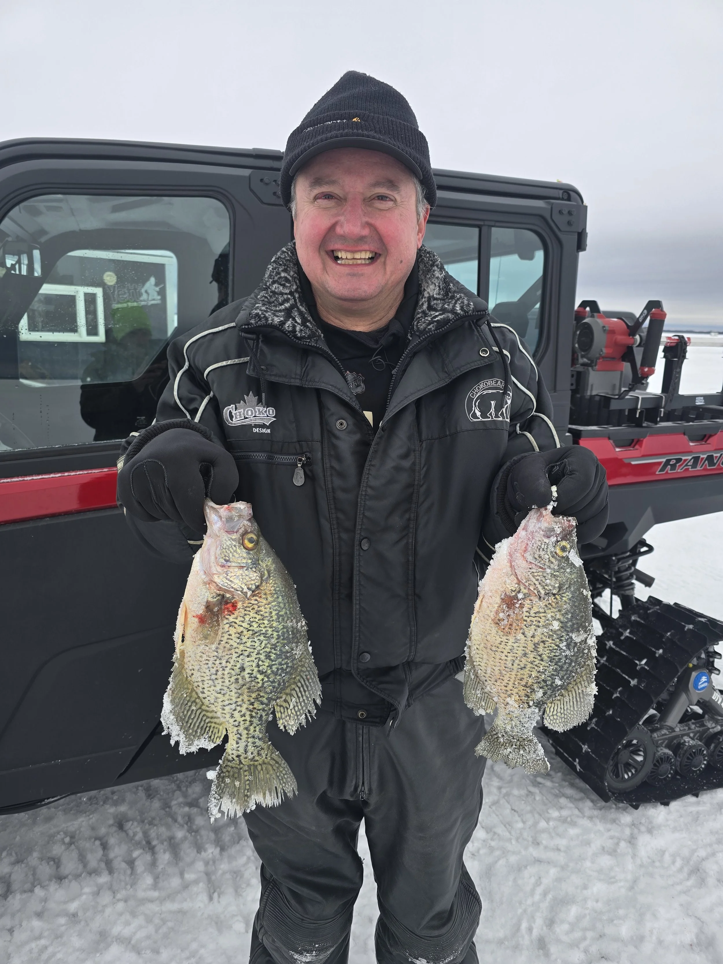 A man smiling and holding two large fish in each hand outdoors on snow, with a black vehicle and equipment in the background.
