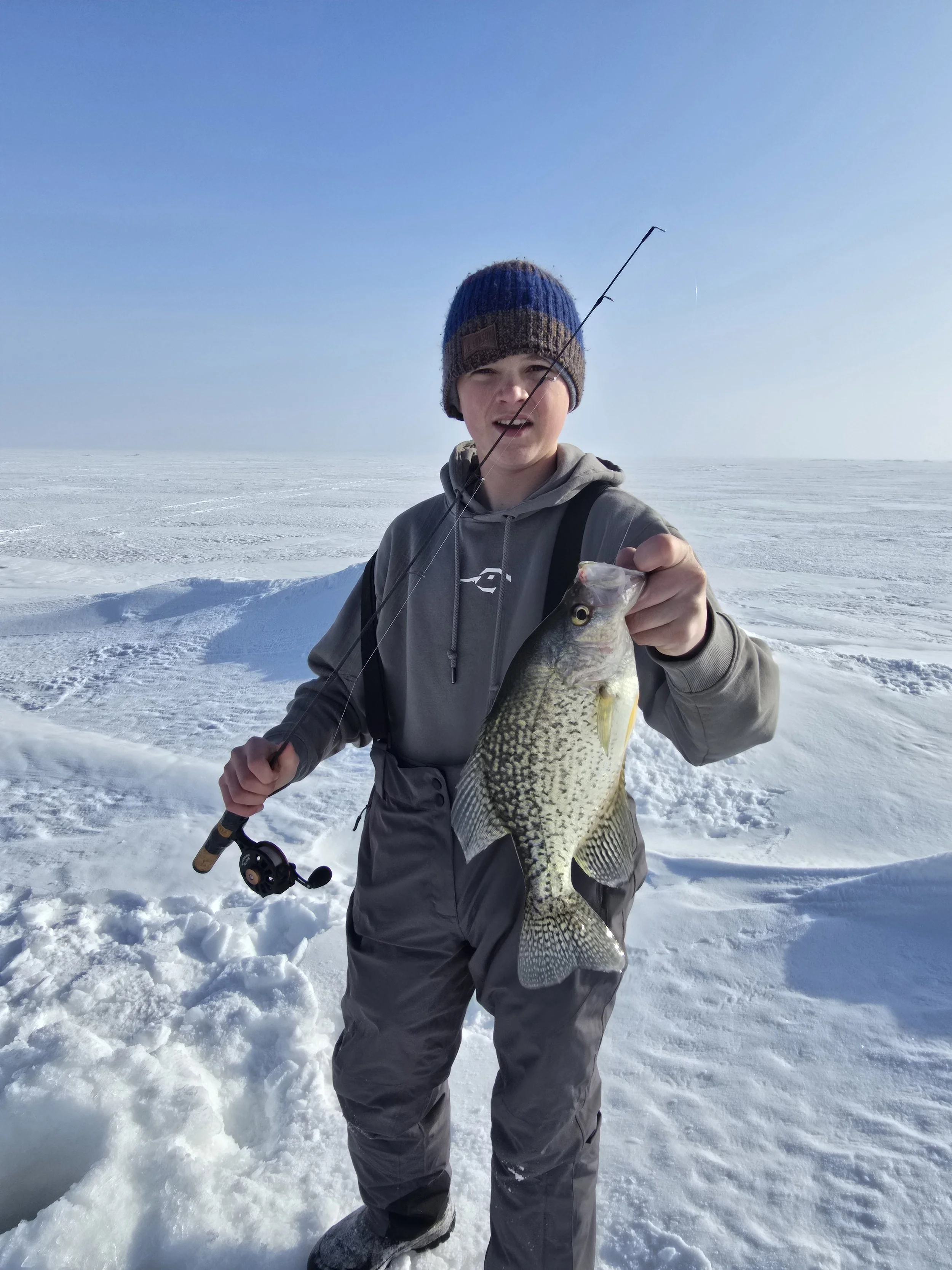 A person standing on snow-covered ice in winter, holding a fish and a fishing rod, wearing a gray jacket, gray snow pants, and a knit hat, with a clear blue sky in the background.