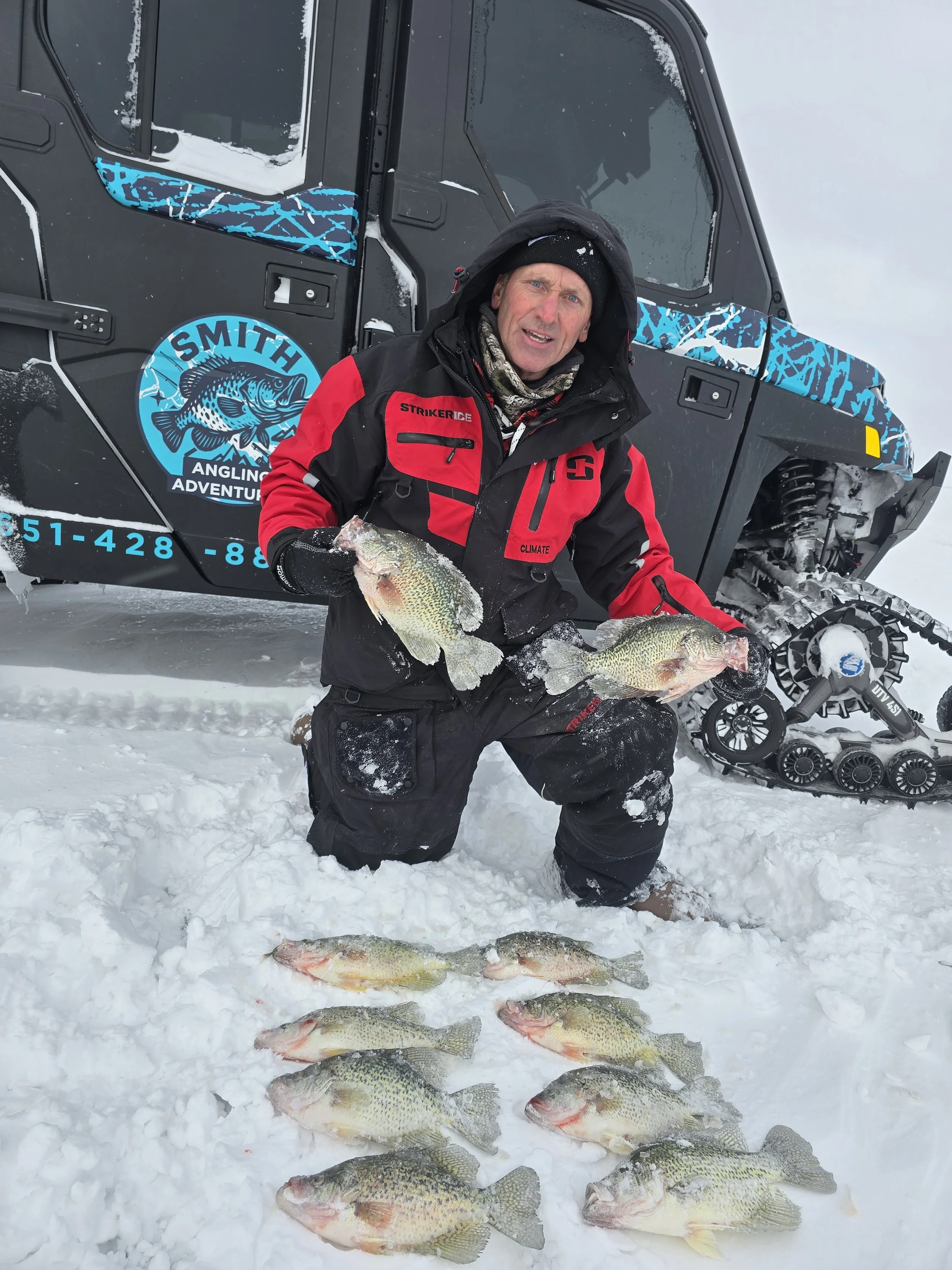 A man in a winter jacket kneeling on snow holding two large fish, with several other fish laid out on the snow in front of him, and a snow-covered off-road vehicle with a logo and contact information visible in the background.