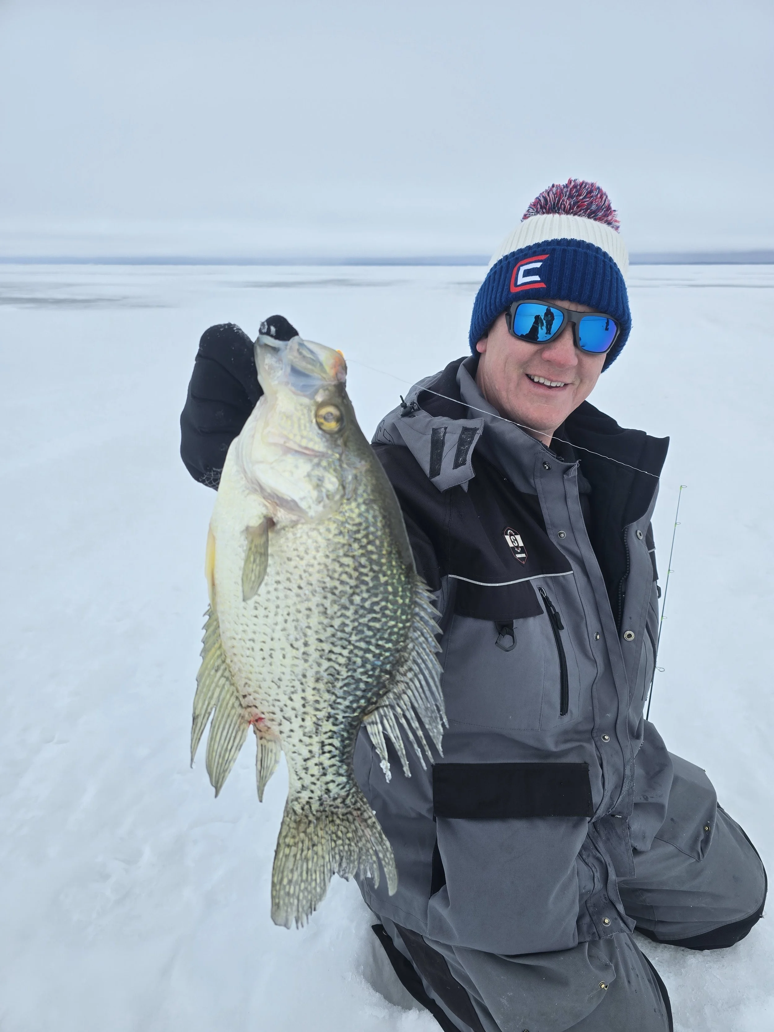 Man in winter clothing and sunglasses holding a large fish on a snow-covered frozen lake.