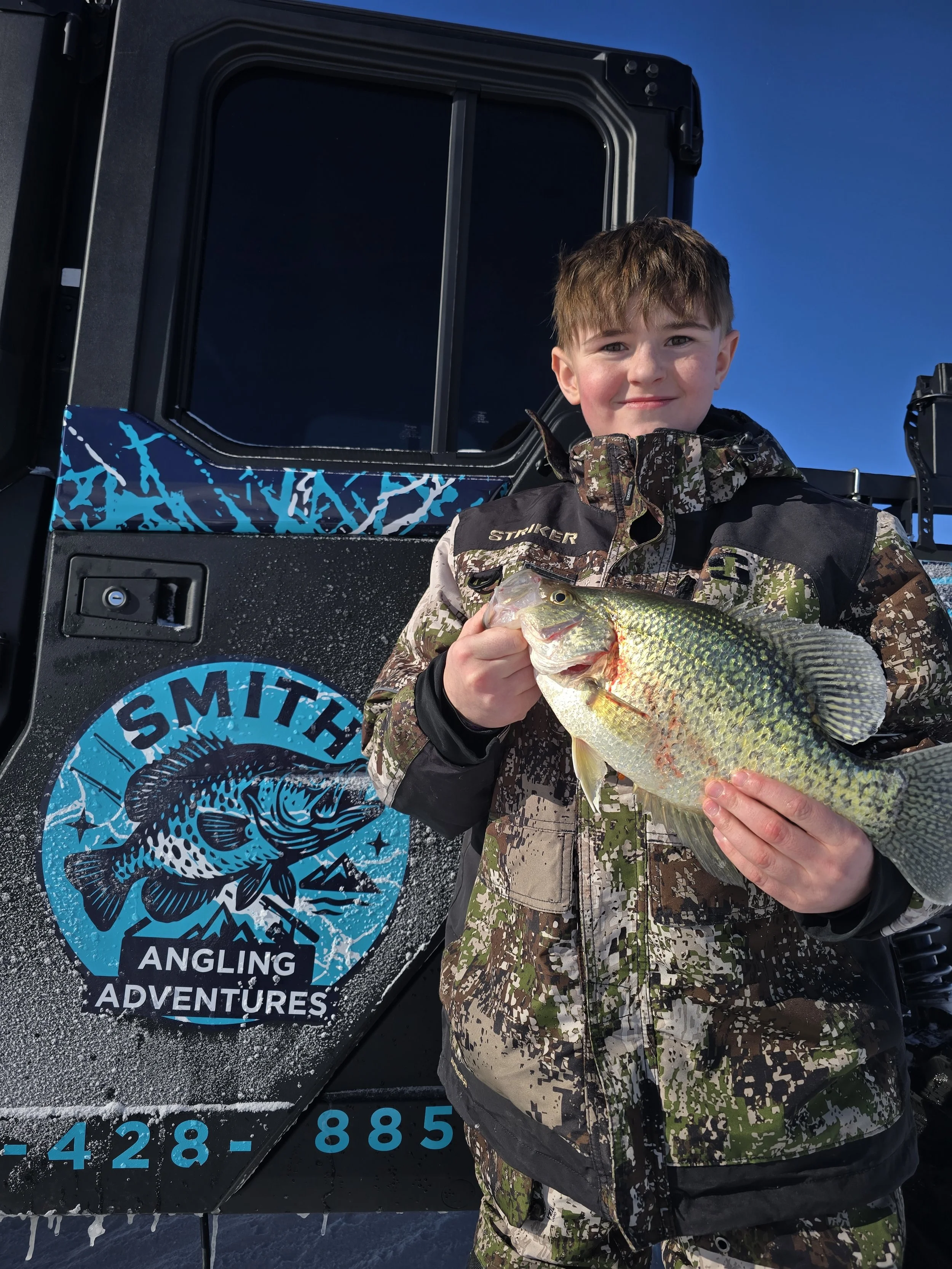 A young boy in camouflage clothing holding a large fish in front of a black vehicle with a blue sticker that reads "Smite Angling Adventures" and other contact information.