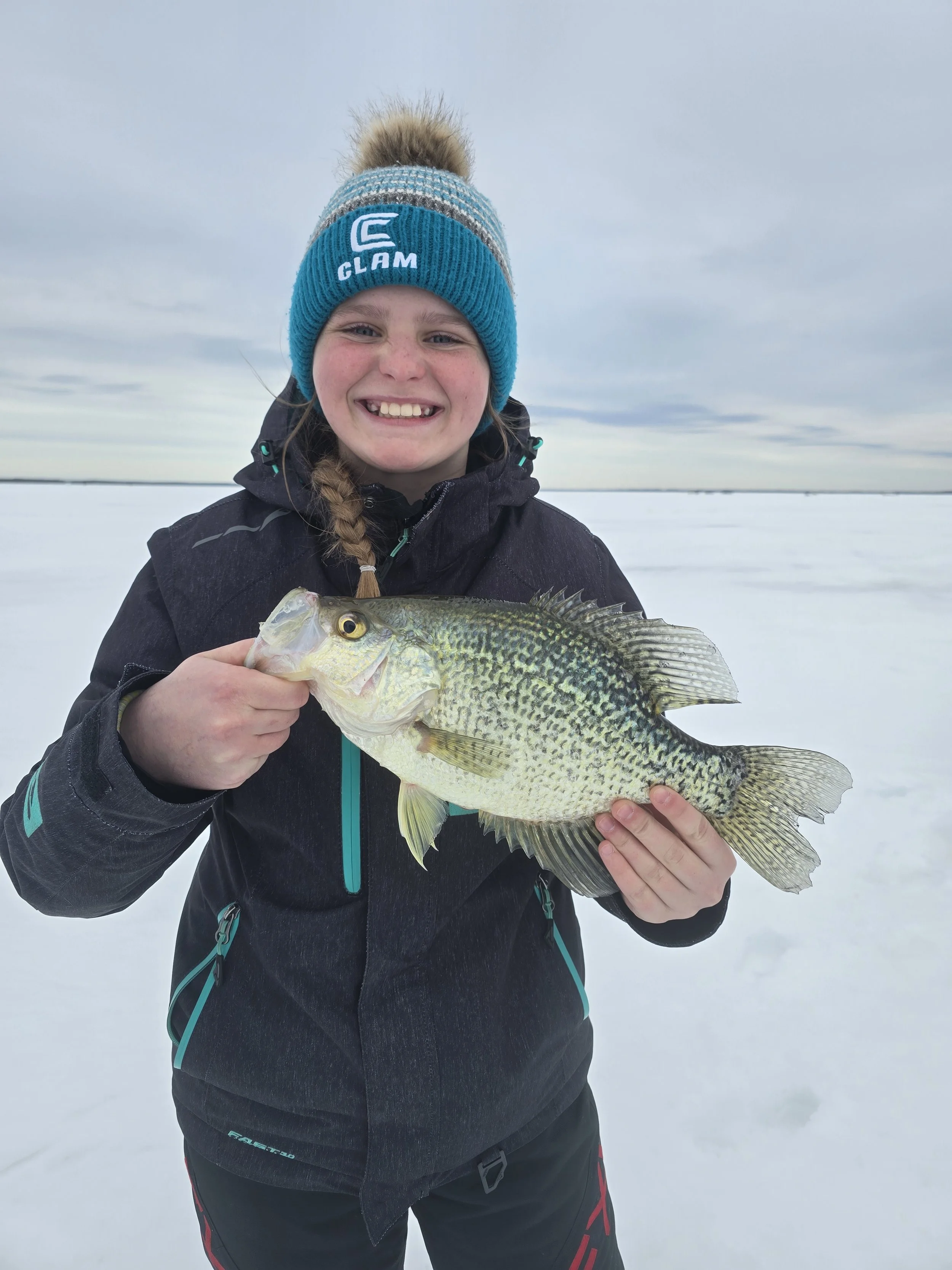 A young girl outdoors in winter clothing holding a fish with a snowy landscape and cloudy sky in the background.
