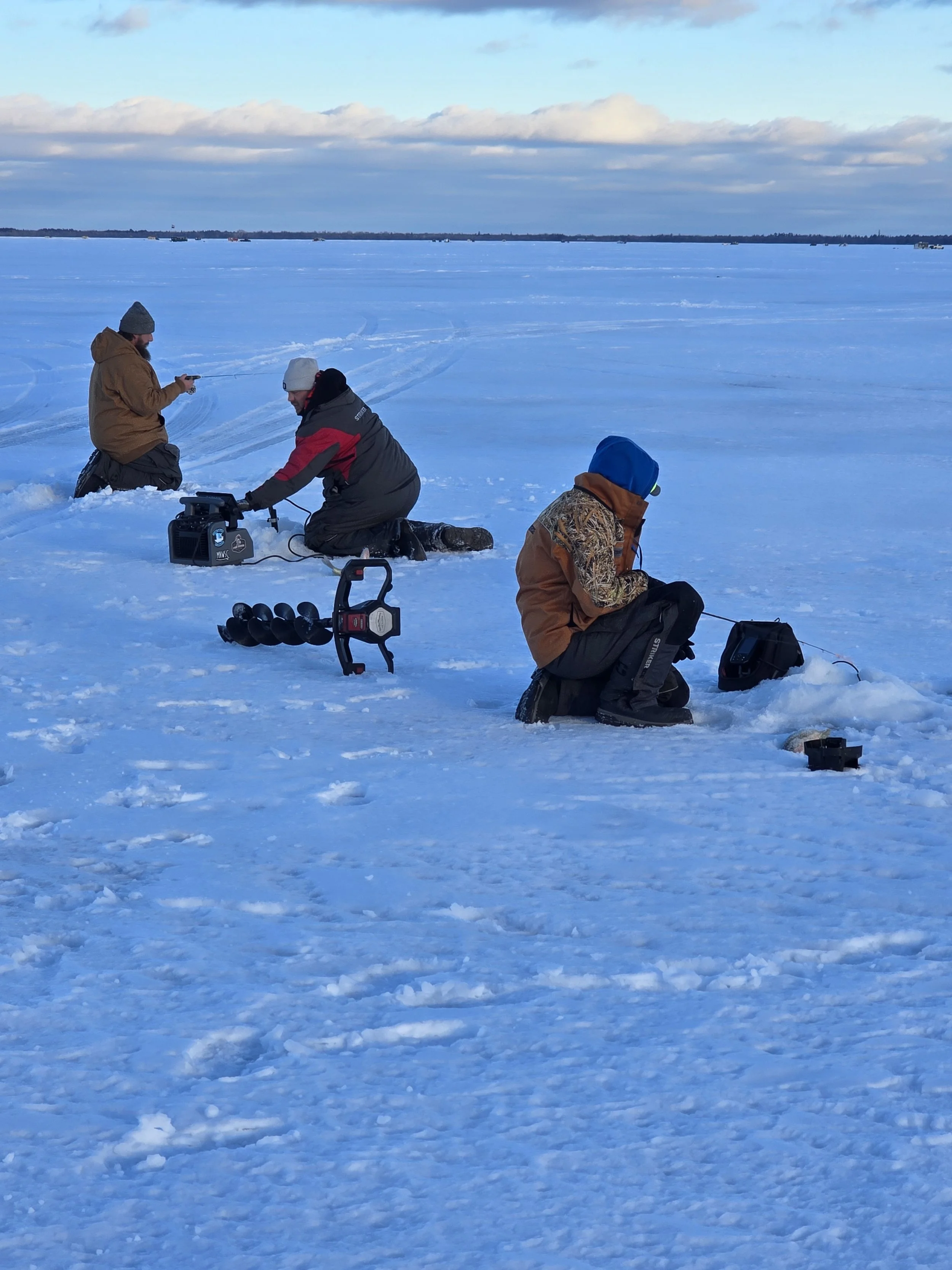 Three people Ice fishing on a frozen Lake. Two are kneeling and one is sitting, dressed in winter clothing and surrounded by fishing equipment.