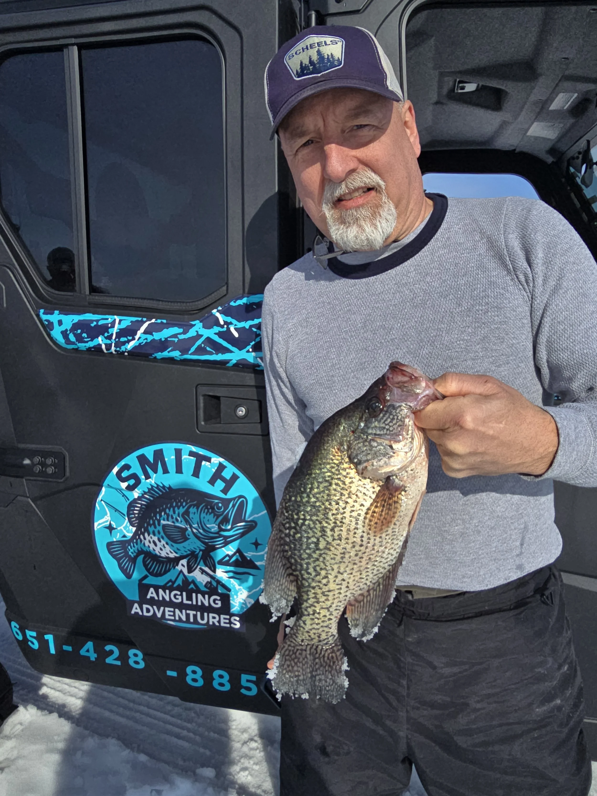 A man with a white beard wearing a purple and gray cap and gray sweatshirt holding a large fish, standing next to a black vehicle with a blue and white sticker that reads 'Smith Angling Adventures'.