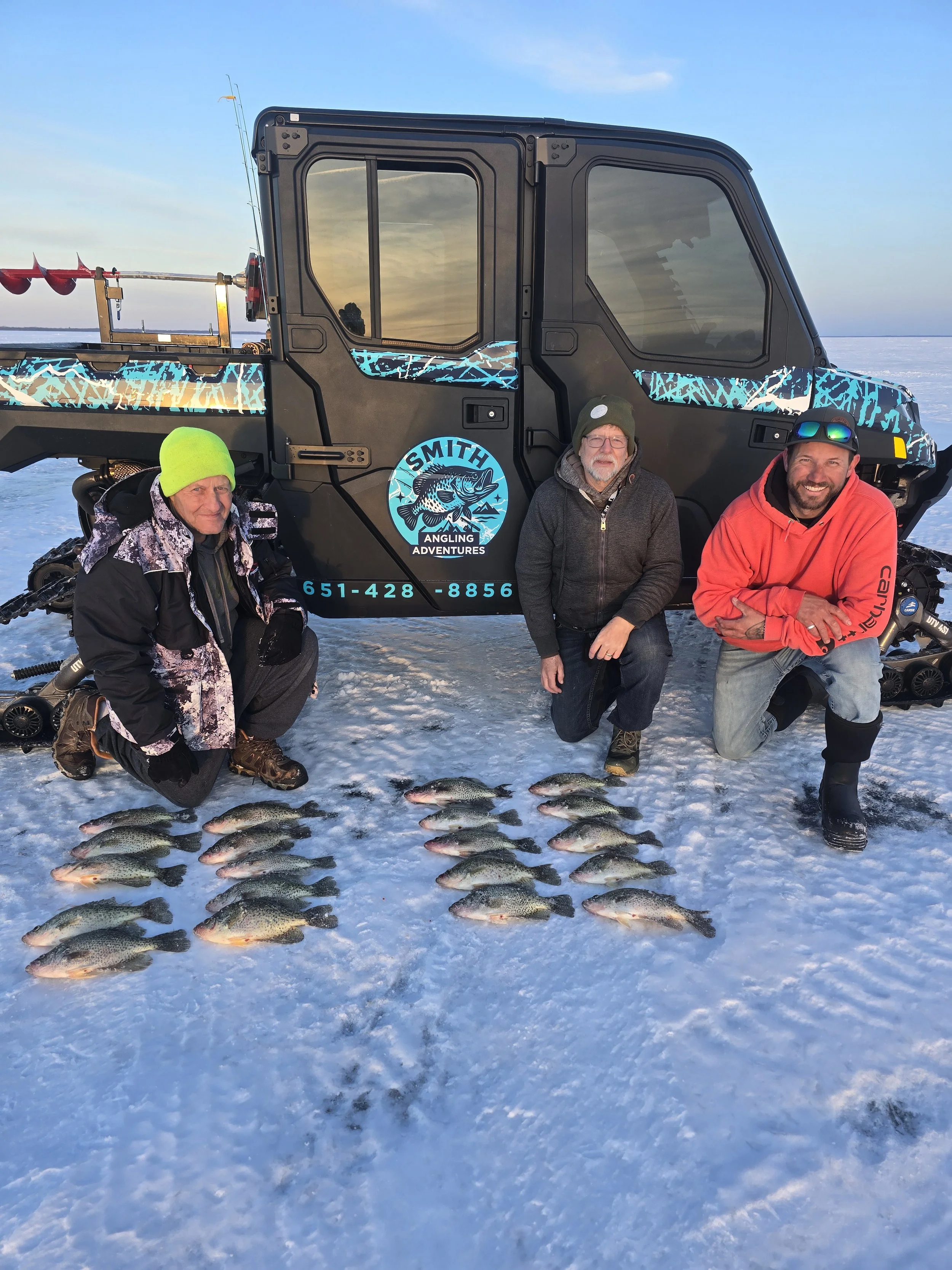 Three men kneeling on snow-covered ice in front of a black vehicle with a blue fish logo, displaying caught fish laid out in rows, with a winter landscape and partly cloudy sky in the background.
