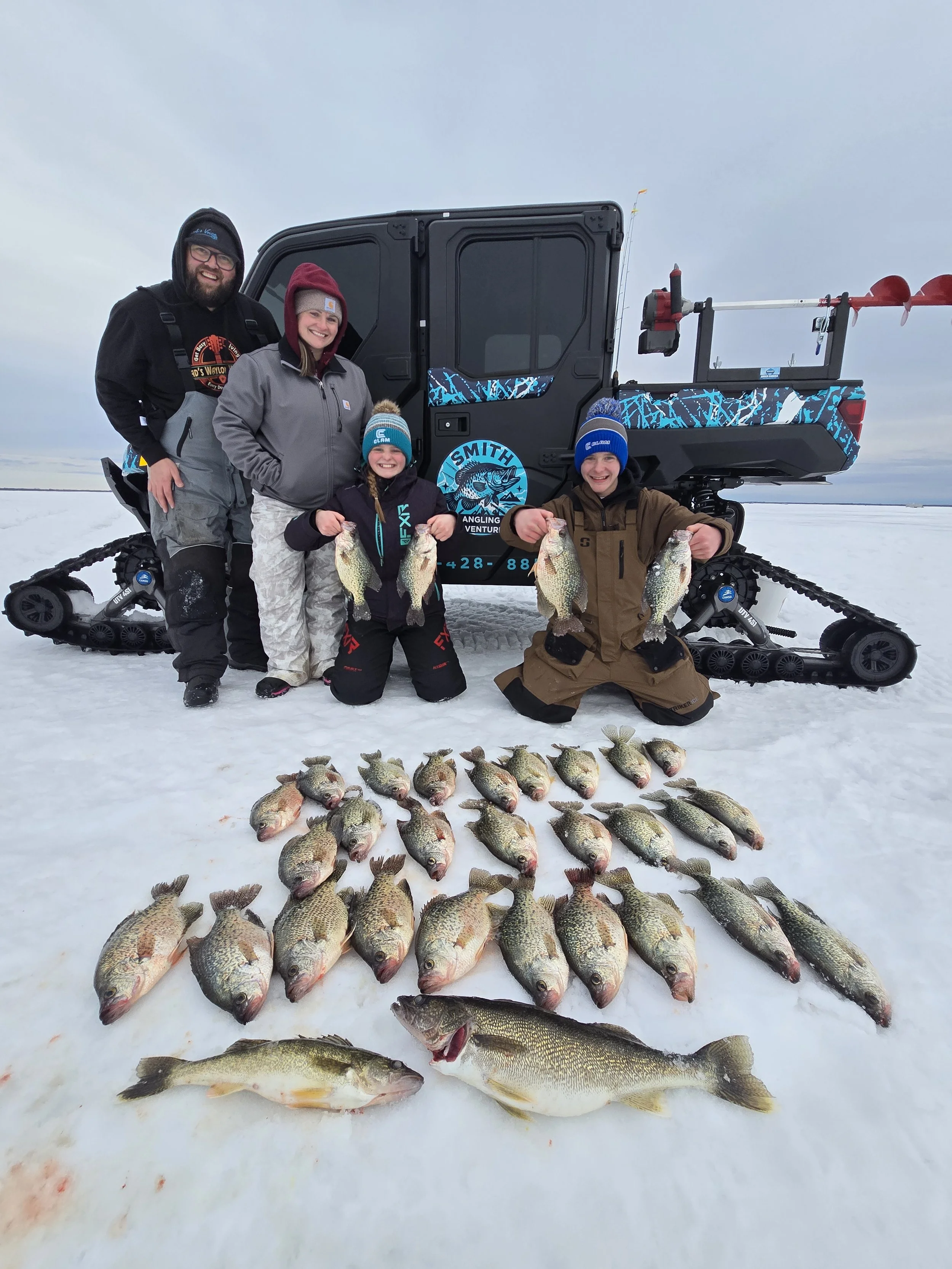 A group of five people, two adults and three children, standing on snow, holding fish they caught during a winter fishing trip with a snow-covered landscape in the background. There's a black utility vehicle with gear, and the group is smiling.