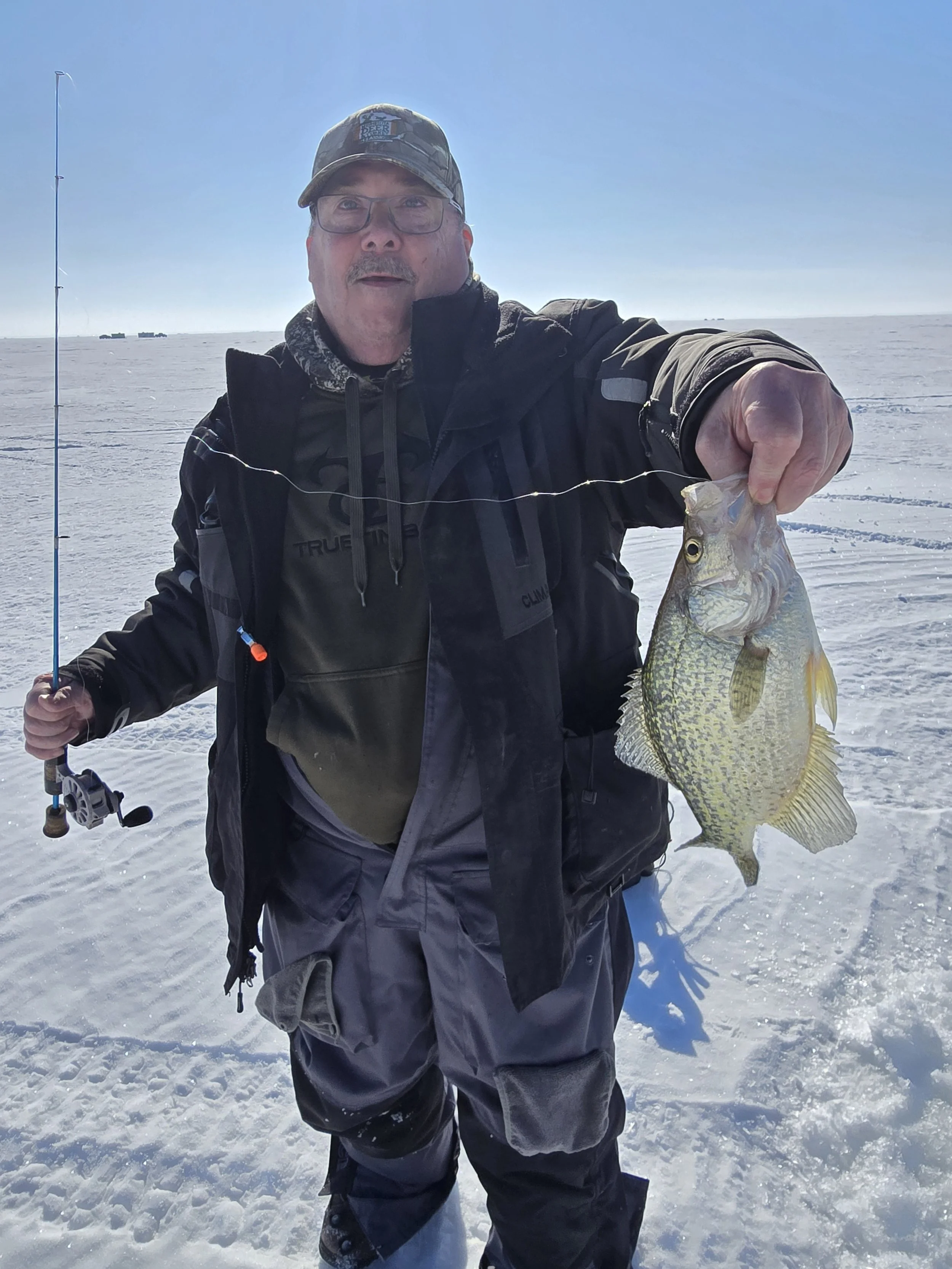 Man dressed in winter clothing holding a fish on a snowy, icy landscape with a bright blue sky.