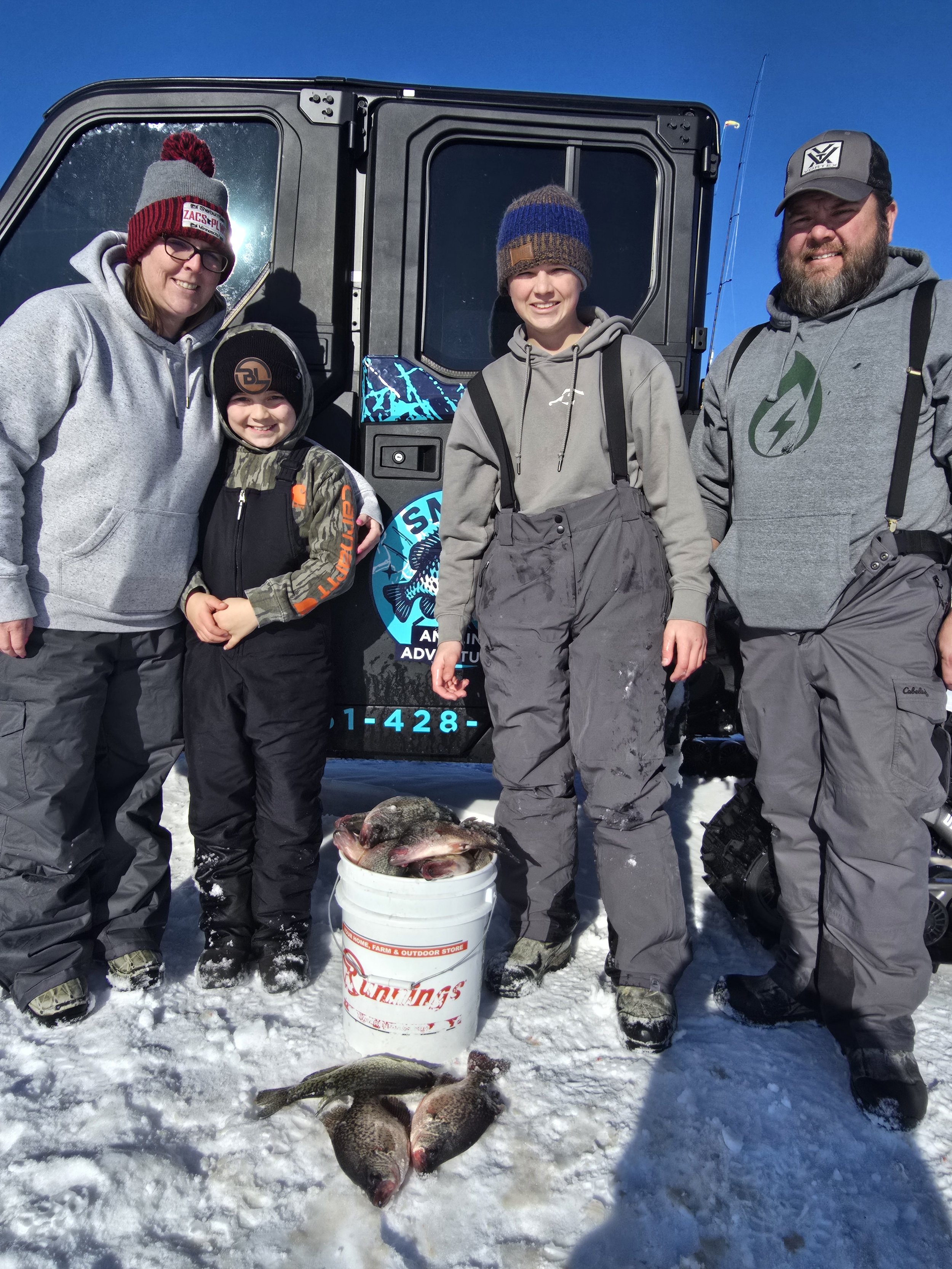 A group of five people, including children and adults, dressed in winter clothing, standing on snow-covered ground, posing with a bucket filled with fish, likely caught while ice fishing. There are large fish on the snow in front of them and a black 