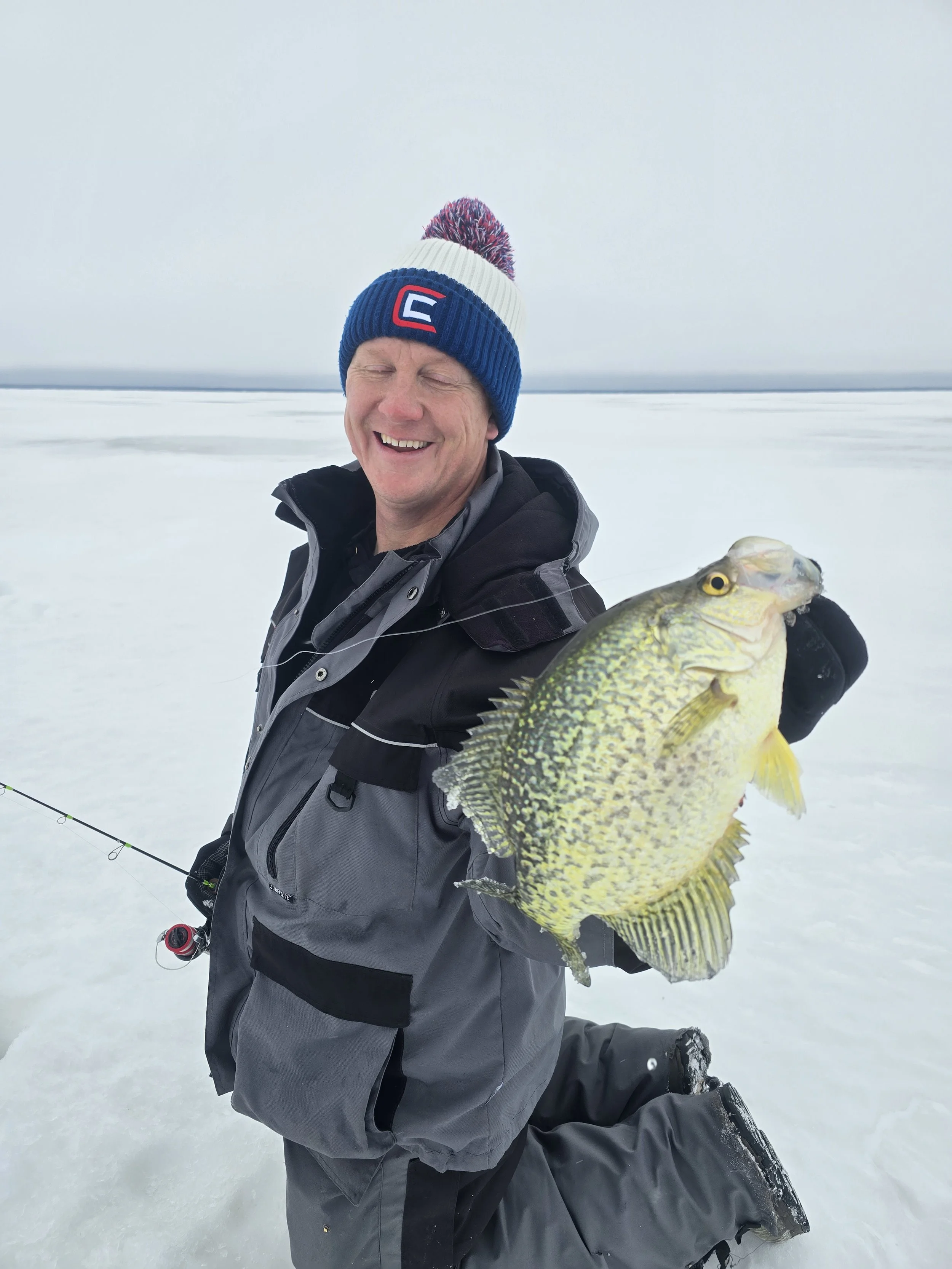 A man kneeling on snow ice outdoors, smiling, holding a large fish, wearing a winter jacket and a colorful knit hat.