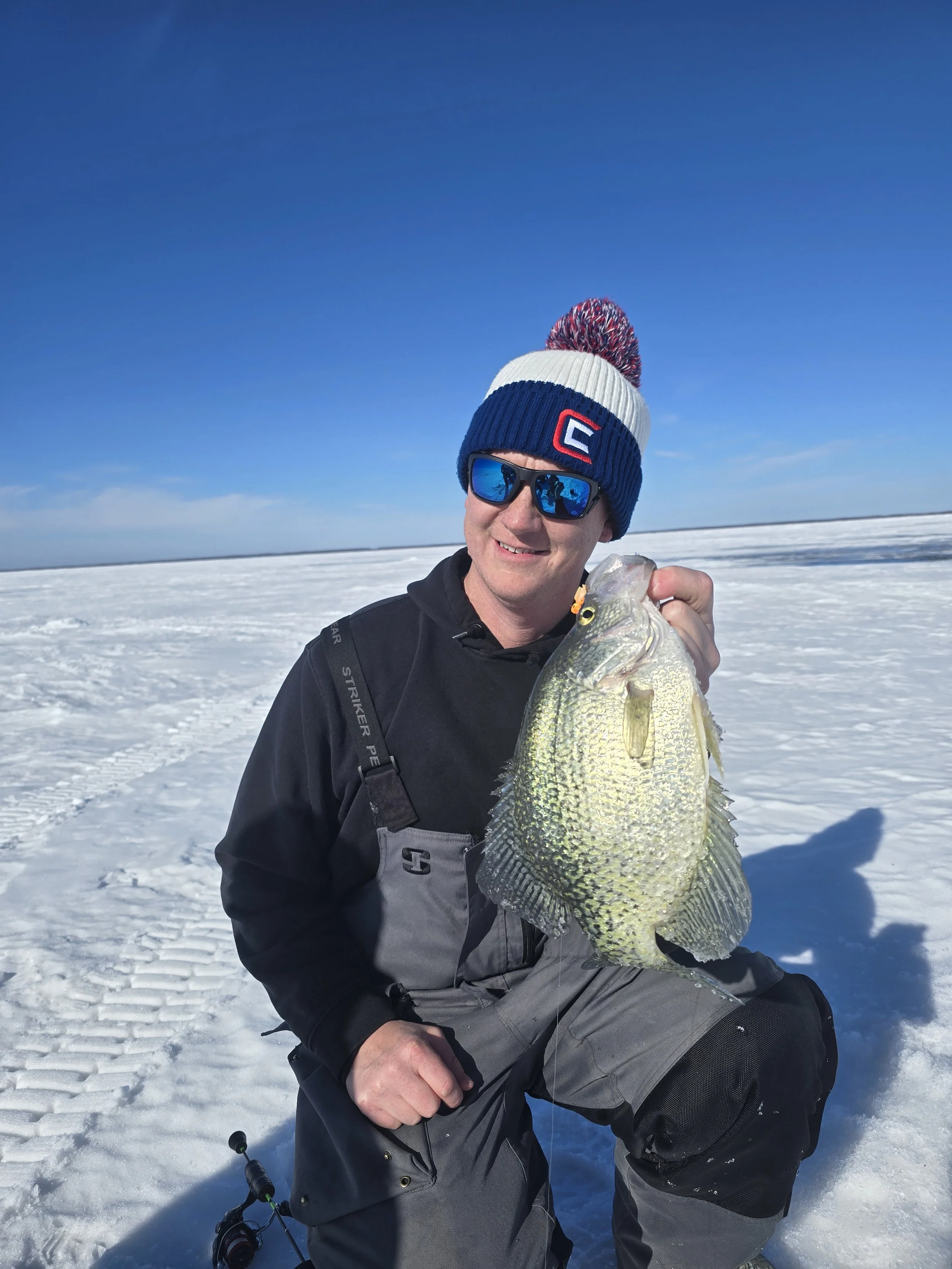 A man in winter clothes and sunglasses holding a large fish, sitting on snow-covered ice with a clear blue sky in the background.