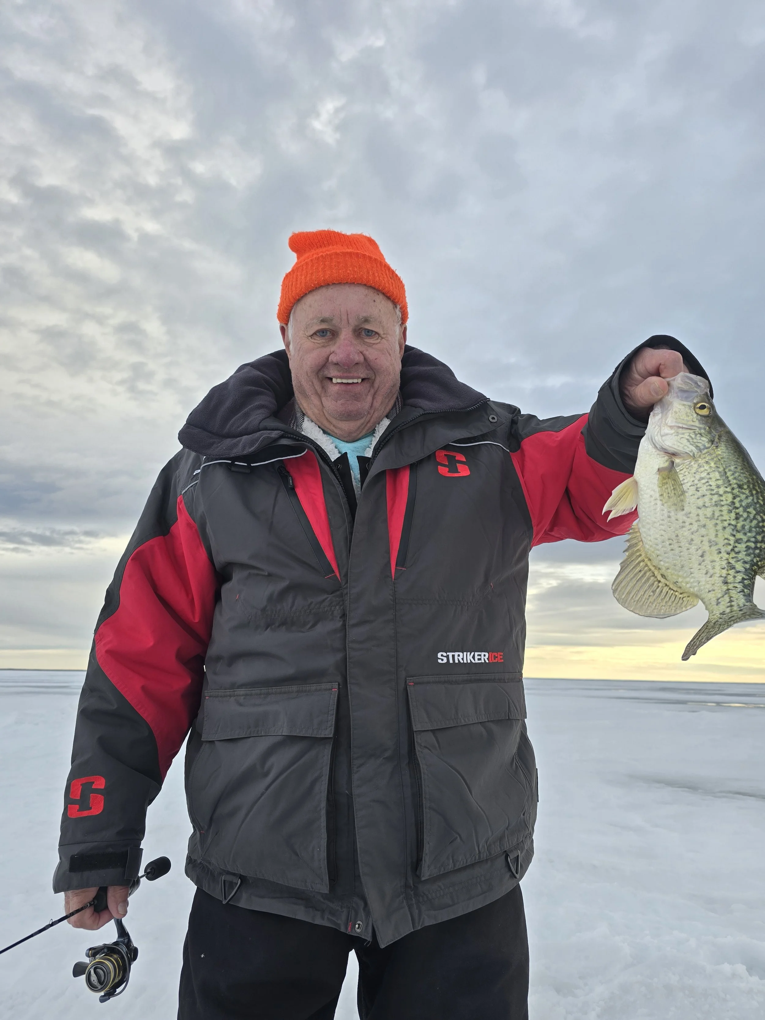 Man wearing a black and red jacket and an orange beanie hat, holding a fishing rod, standing on a snowy landscape during cloudy weather, displaying a large fish.